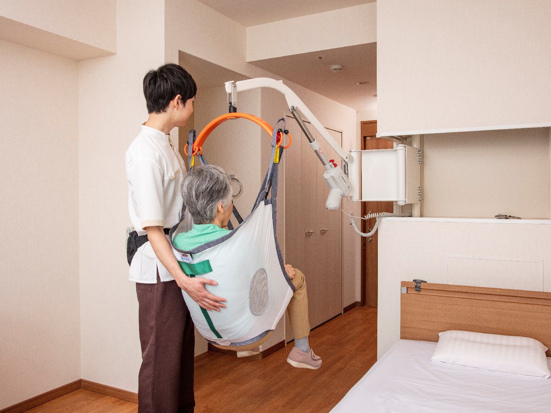 吊り下げてベッドまで運べるリフトに乗っている白髪交じりの高齢者とそれを支えている介助のスタッフ