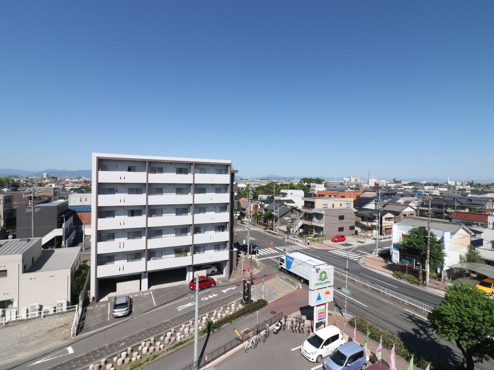 マンションのような白い建物が正面に建っている。その右横には4車線の広い道路があり、1台の普通車と大型トラックが通行中である。