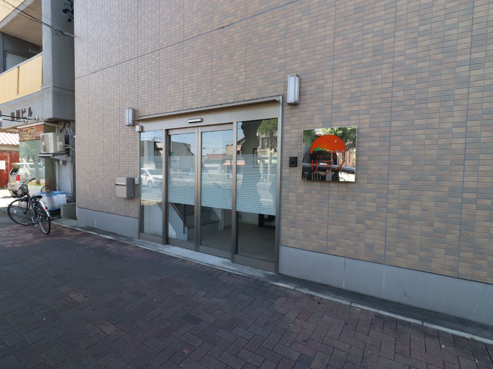 煉瓦調のフラットな壁に4枚のガラスの部分があり、真ん中2枚が自動ドアになっている。横には赤い字で施設名を書いた看板が貼ってある。