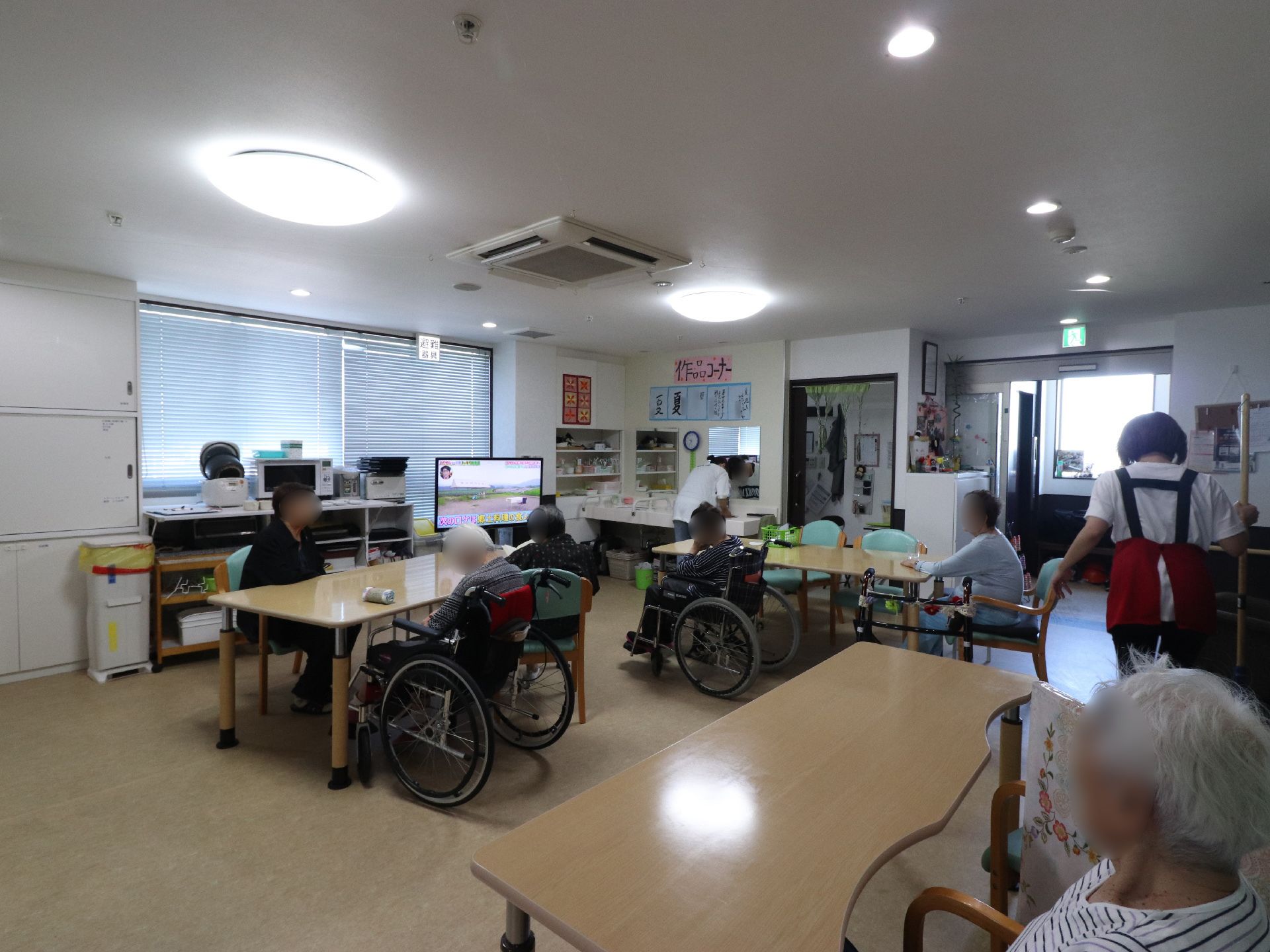 木目調デザインのテーブルの周りに、数人の入居者の方々が座ってテレビを見ている。エプロン姿のスタッフが右端に立っている。