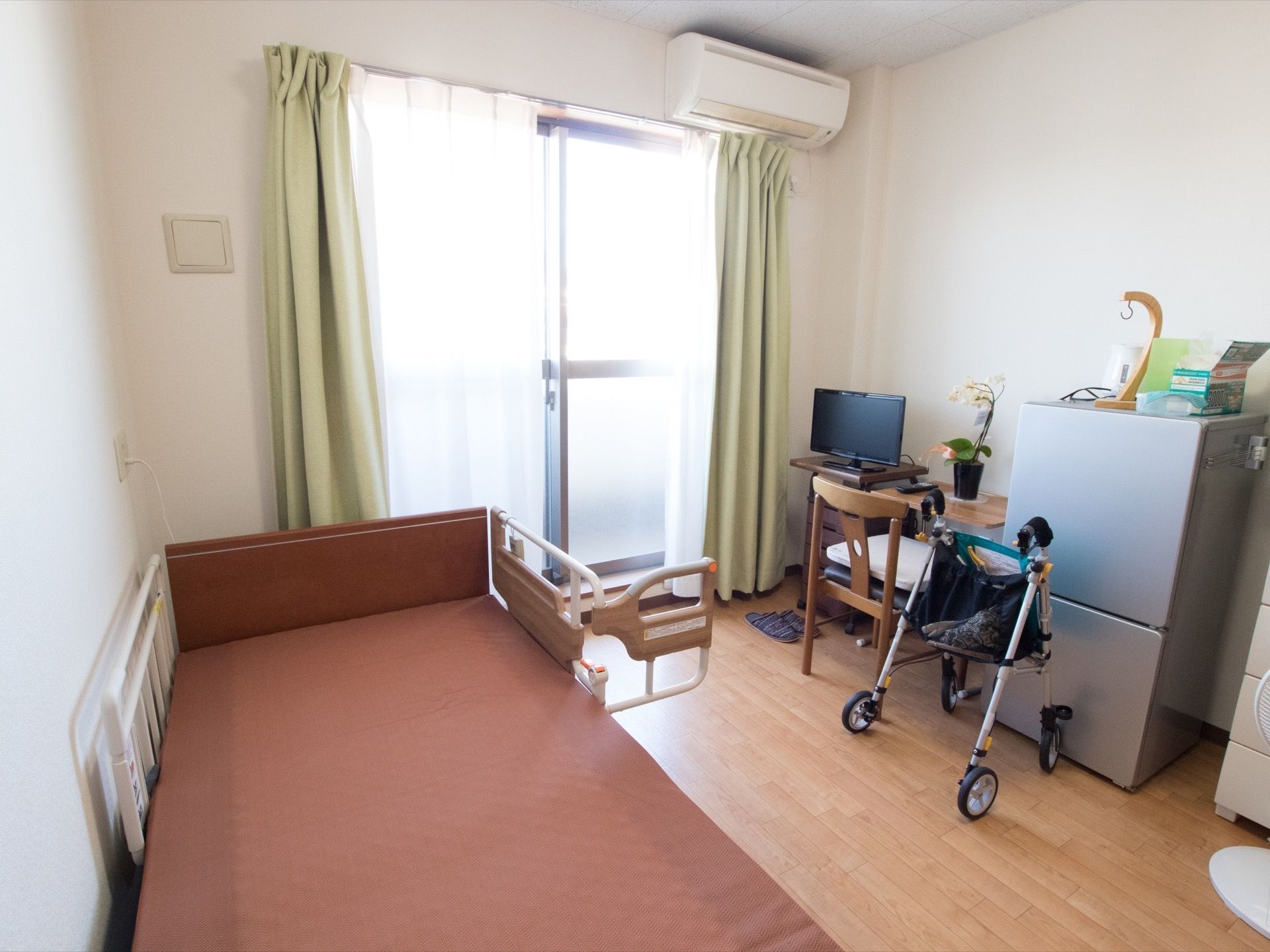 白で統一された明るい日当りのよい居室である。介護用ベッドや冷蔵庫、テレビ、カーテン、机が完備されている。