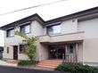 2階建ての施設の真ん中に玄関入口がある。前には階段があり横に木が植わっている。入口の横には2本のシルバーの柱が付いている。