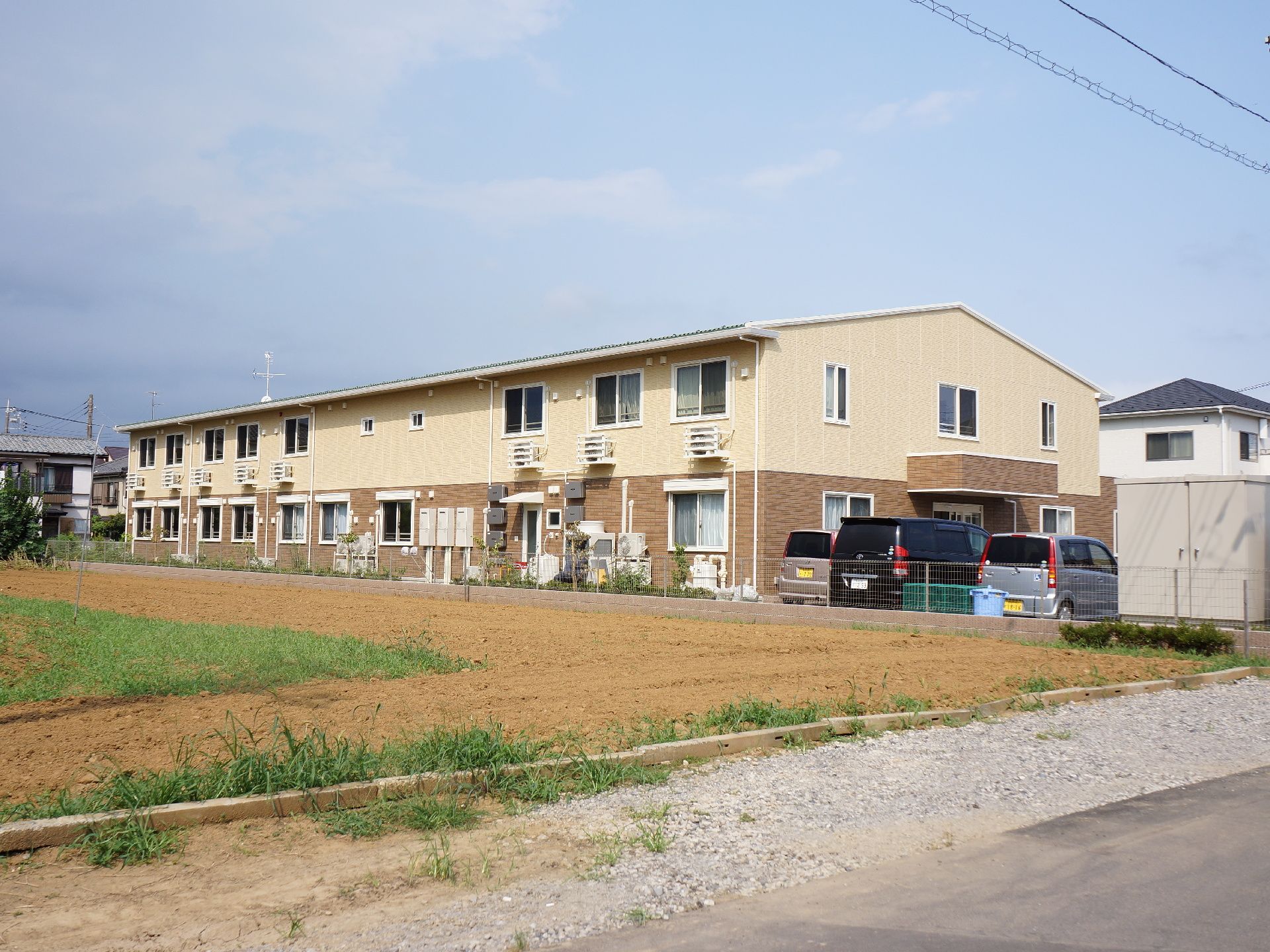 施設外部は大きな畑が面積を占めており、その奥に施設外部が見えている。その横には車が何台か停まっている。