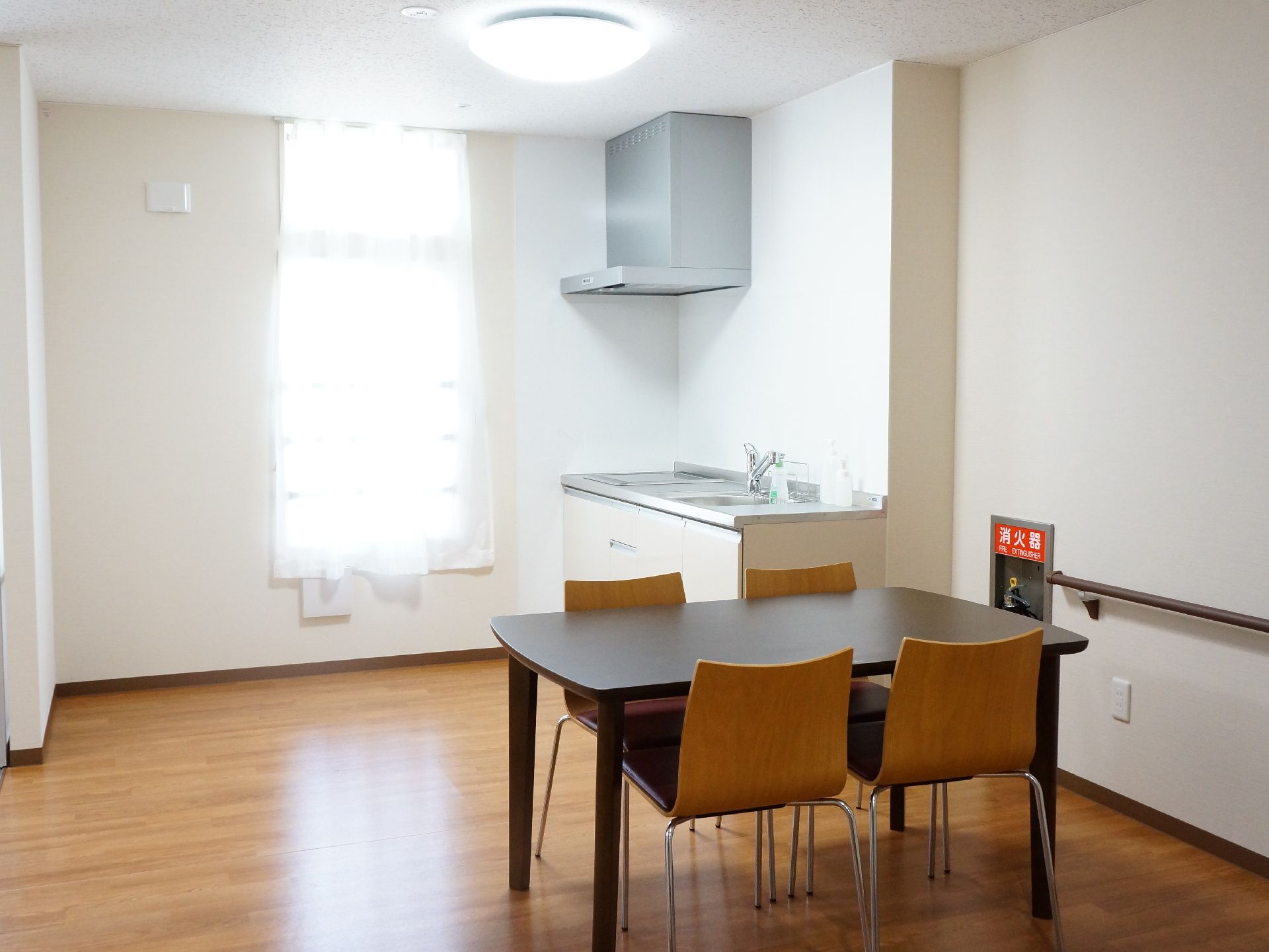 天井の照明が点いた状態となっている部屋には1つのテーブルとイスが置いてある。壁際にはキッチンと消火器の設備がある。