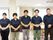 5人の職員が横一列になって並んでいる。左側から順に女性が2人おり、男性が3人いる。全員同じ紺色の上着を着ている。