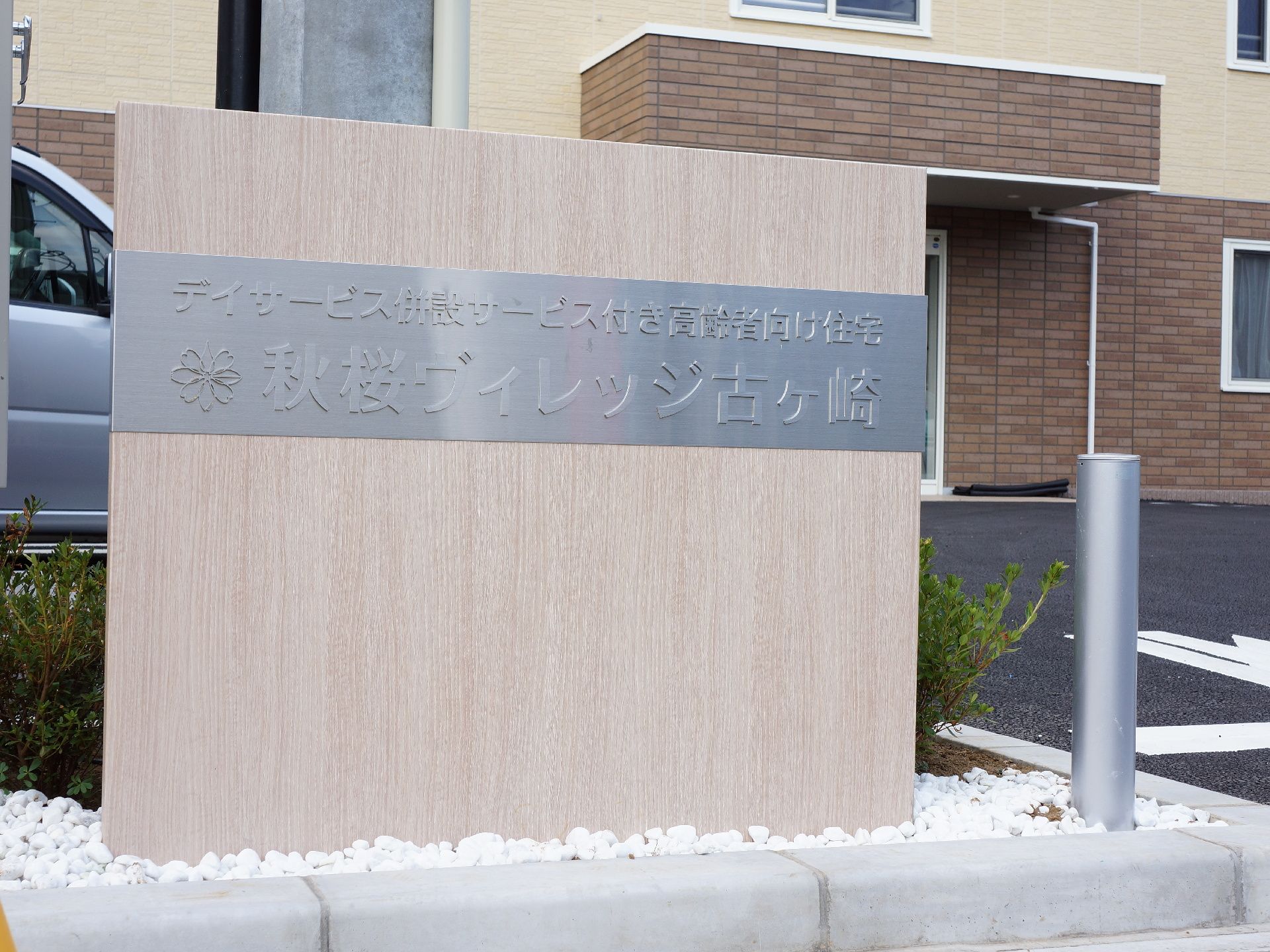 正面に施設名が書かれた表札が立っている。文字部分の周りは銀色になっており、それ以外は白色の木目調になっている。右側にはポールが立っている。