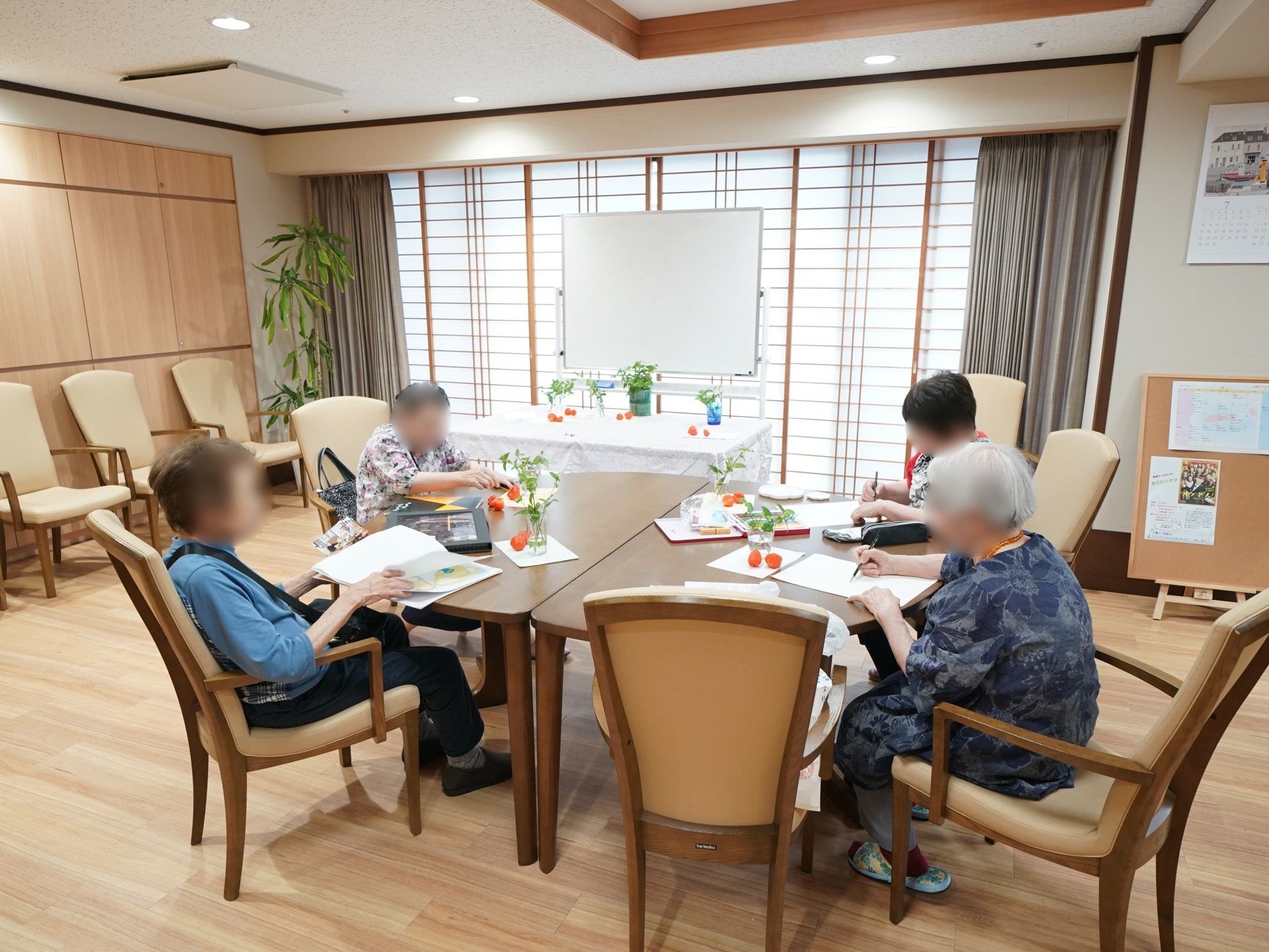 共有スペースでは入居者様がレクリエーションをしている。障子越しに日差しが差し込んでいる。ホワイトボードが置かれている。