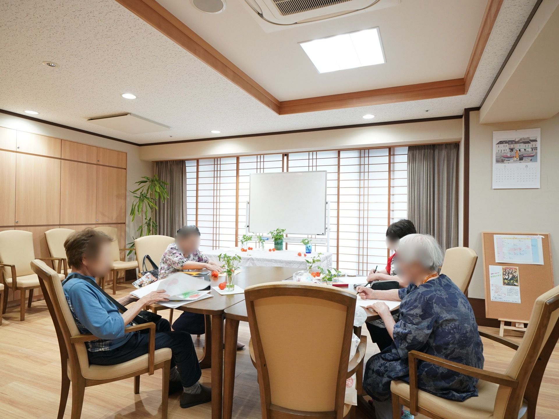 障子のある和風の部屋にはボードがあり、レクリエーションの案内や行事予定が掲示されている。テーブルでは４人の入居者様が絵を描かれている。