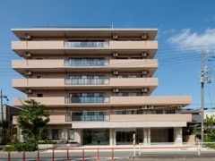 ベージュ色の外壁が特徴である施設が建っている。施設のそばには木が植えられており、緑を感じることができる。