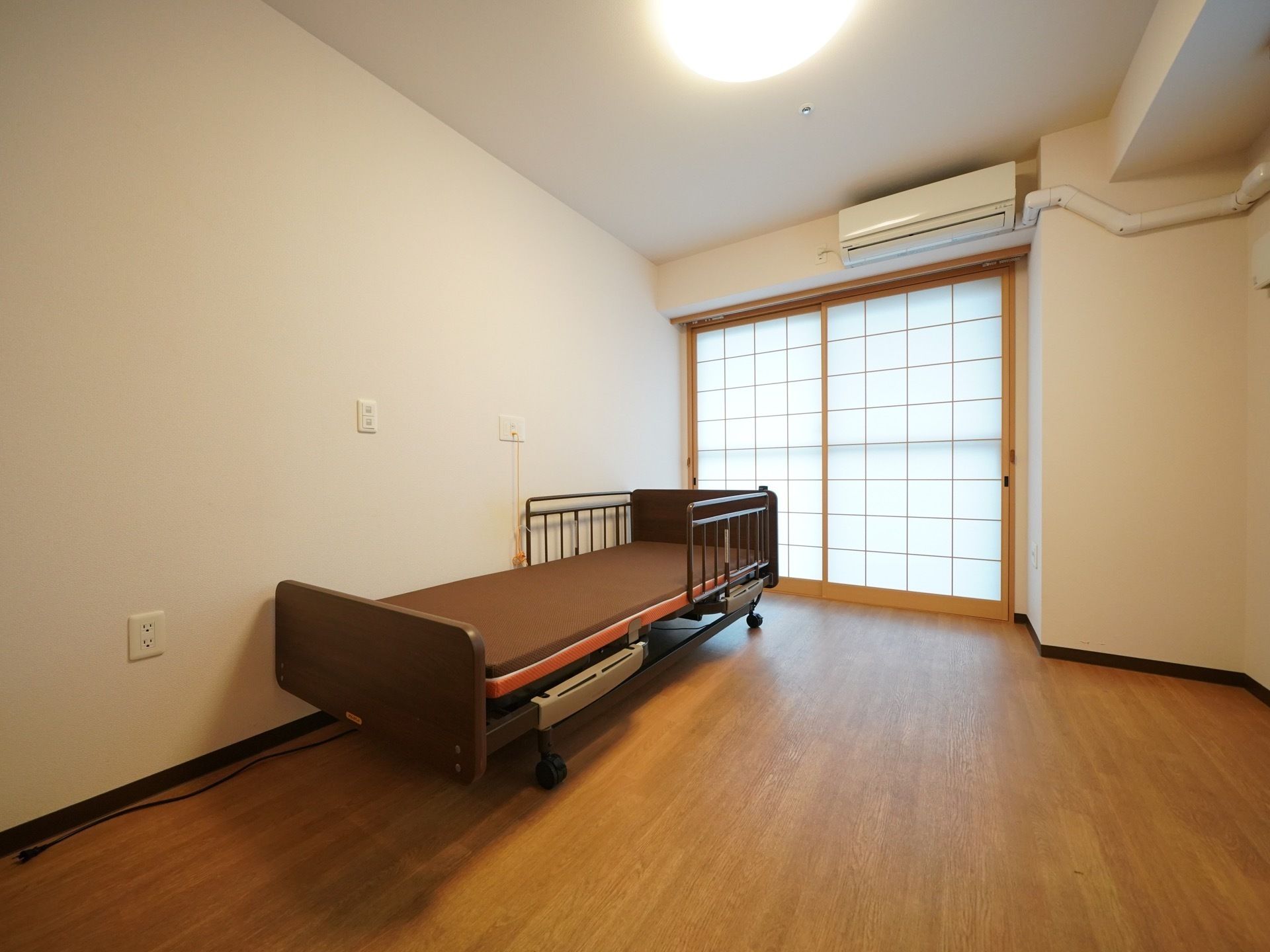 エアコンが完備された個室タイプの居室である。キャスターとフェンスのついた介護ベッドが壁沿いに置かれている。