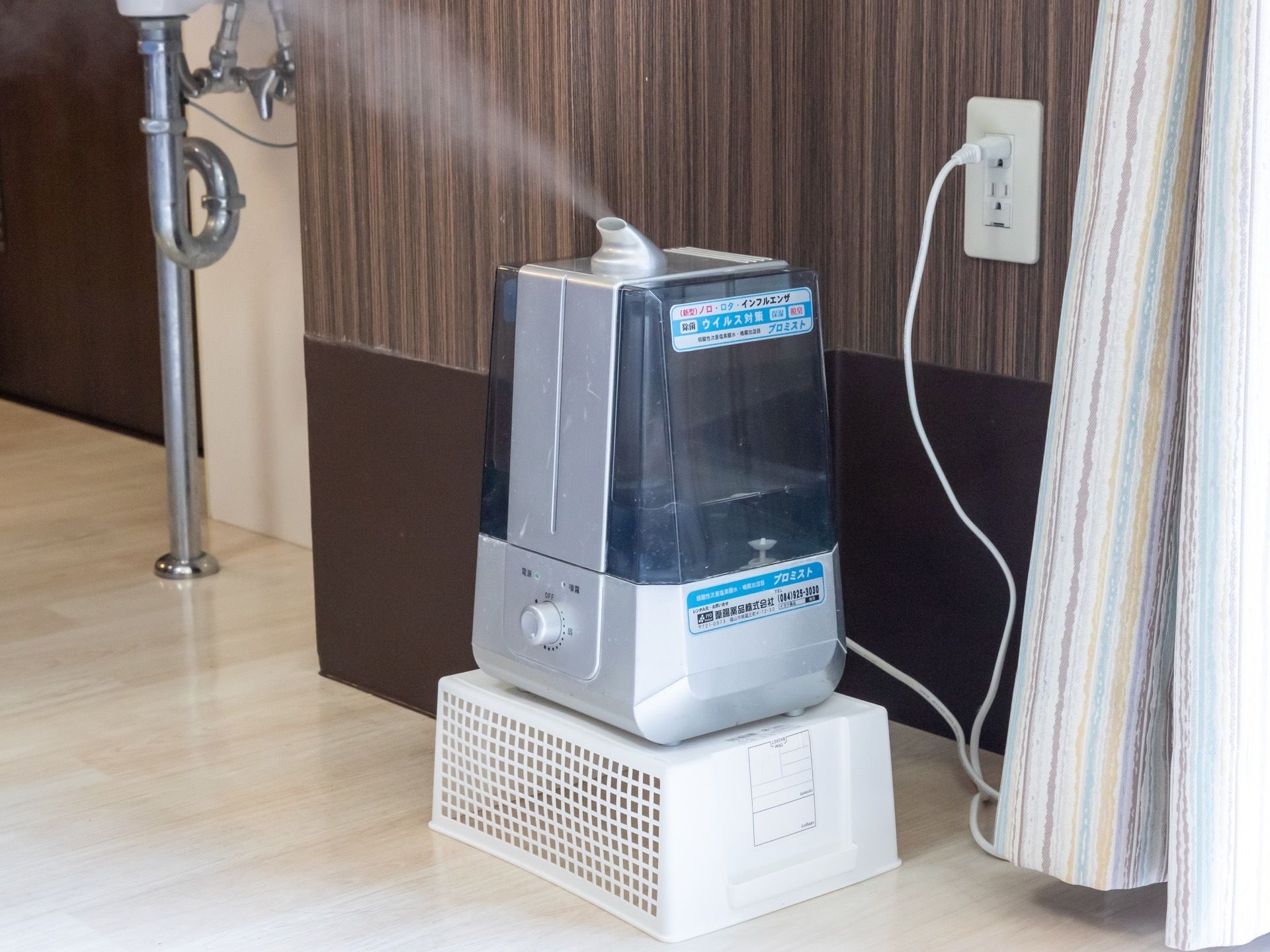 茶色の壁のコンセントから電源を取り、運転している加湿器である。逆さまにした白いカゴの上に置かれている。