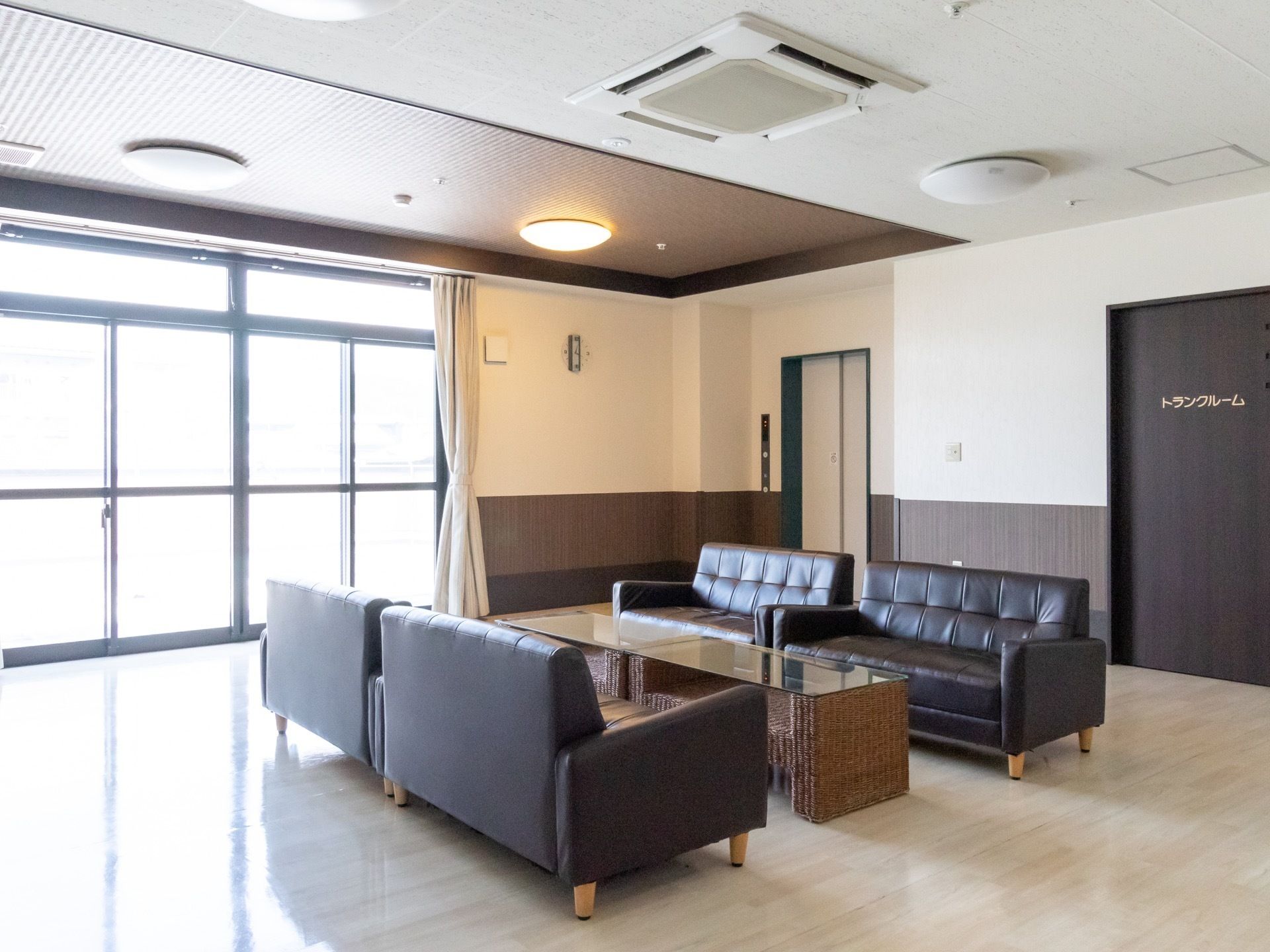 天井には埋め込み式のエアコンが設置され、大きな窓から光が差し込んでいる空間である。白い扉のエレベーターが１基ある。