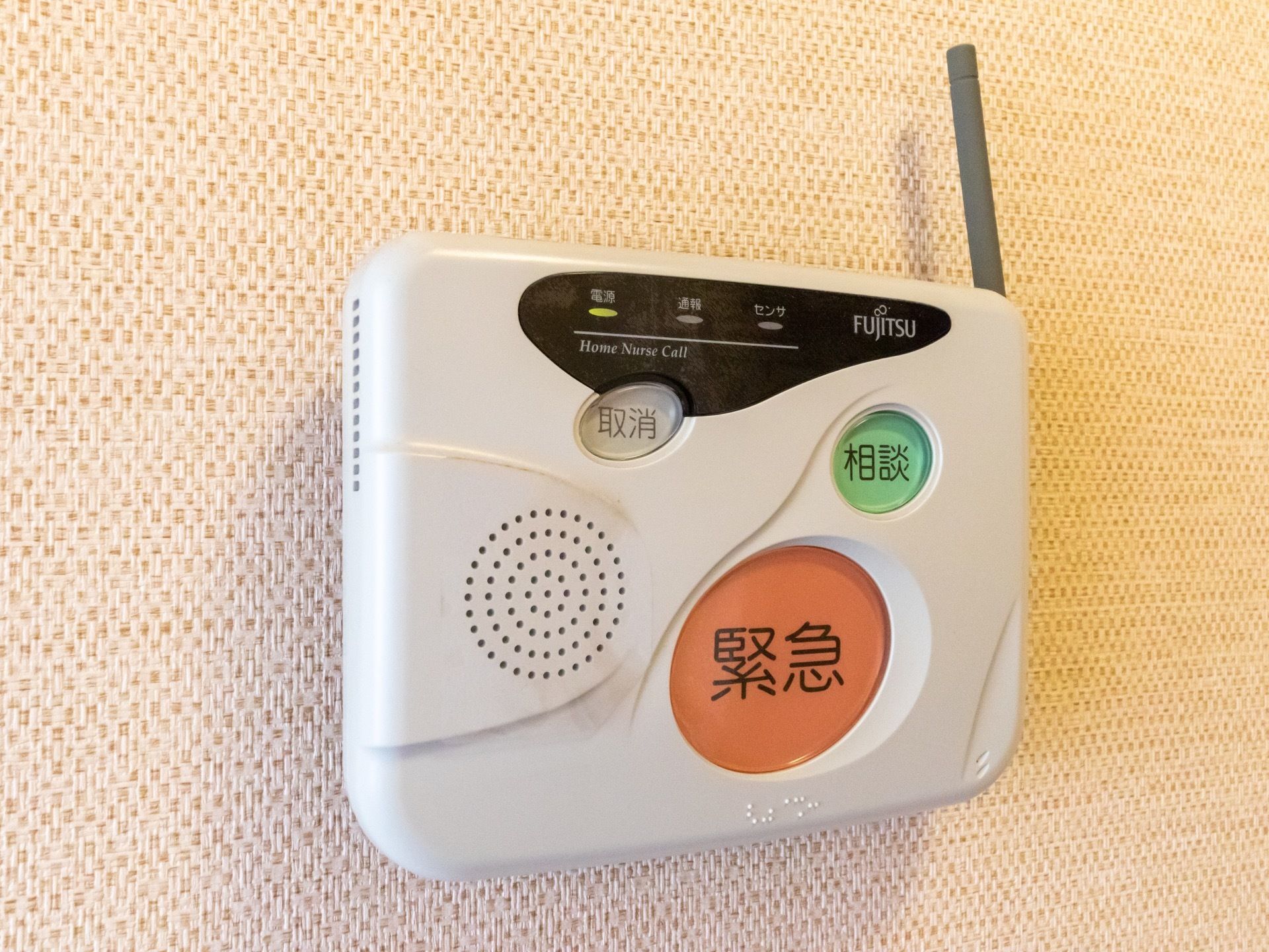 茶系の壁に、ナースコールの機械が取り付けられている。緊急、相談、取り消しの３種類にボタンが前面にあり、側面にはアンテナがついている。