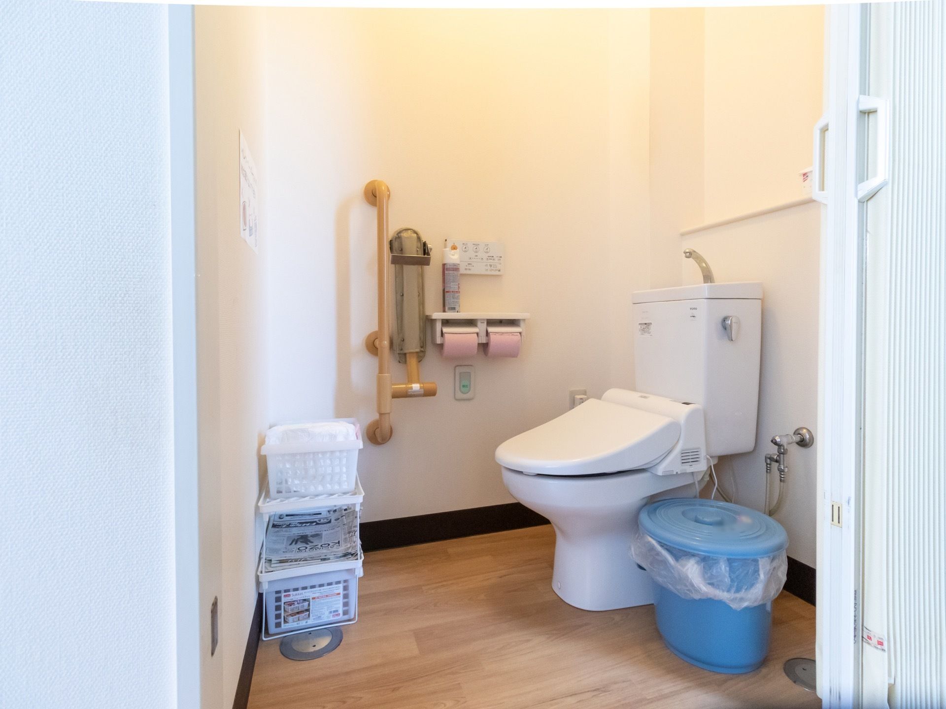 白を基調とした清潔感のあるトイレである。壁には手すりがある。ペパーホルダーにはピンクのトイレットペーパーが取り付けてある。