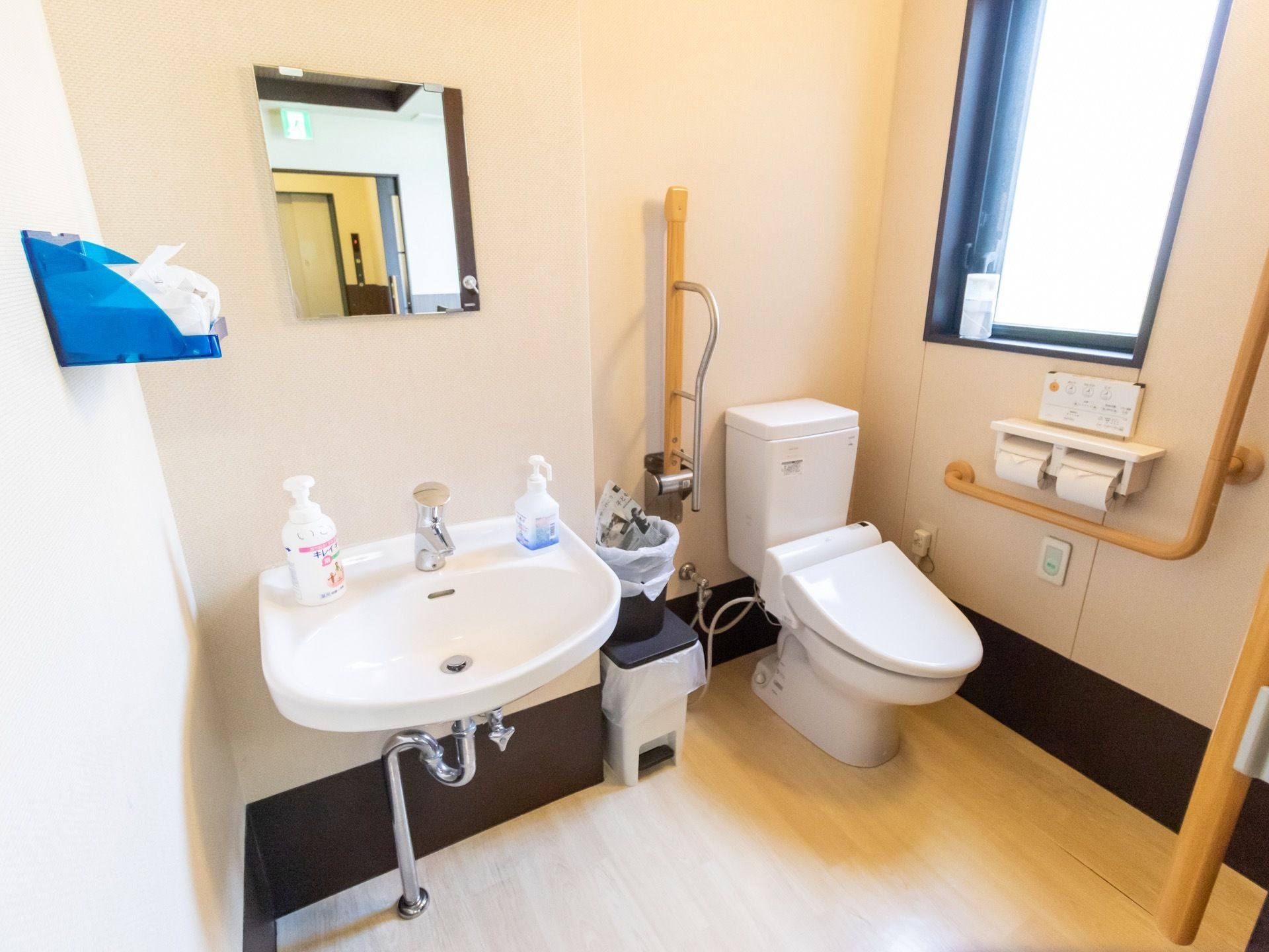 壁に鏡が取り付けられた手洗い場が併設されているトイレである。壁には複数の木製の手すりが設置されている。