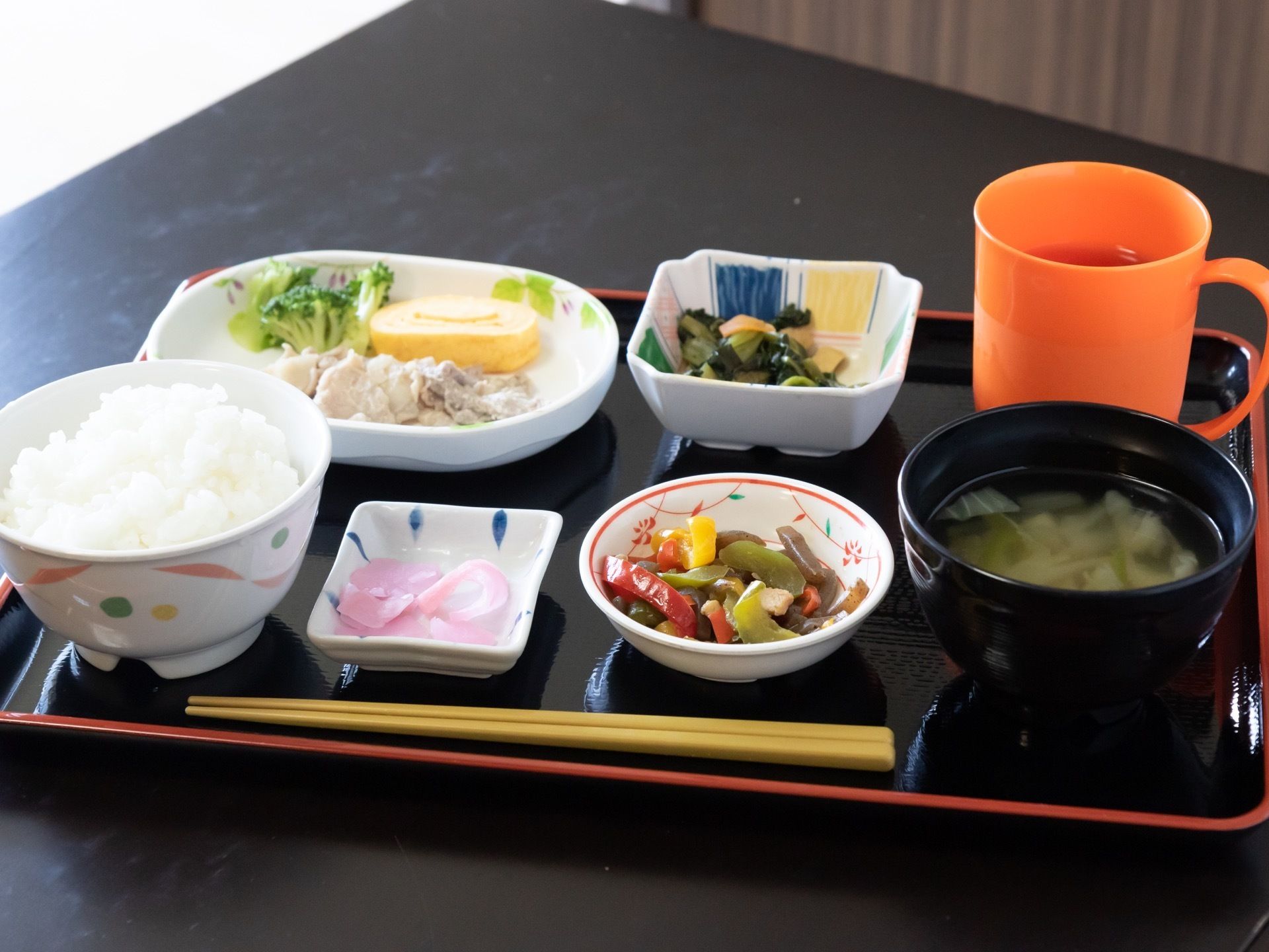黒いテーブルの上にお盆にがあり、その上に１人分の食事が並べられている。主菜のほかに小鉢が２つあり、お茶も置かれている。