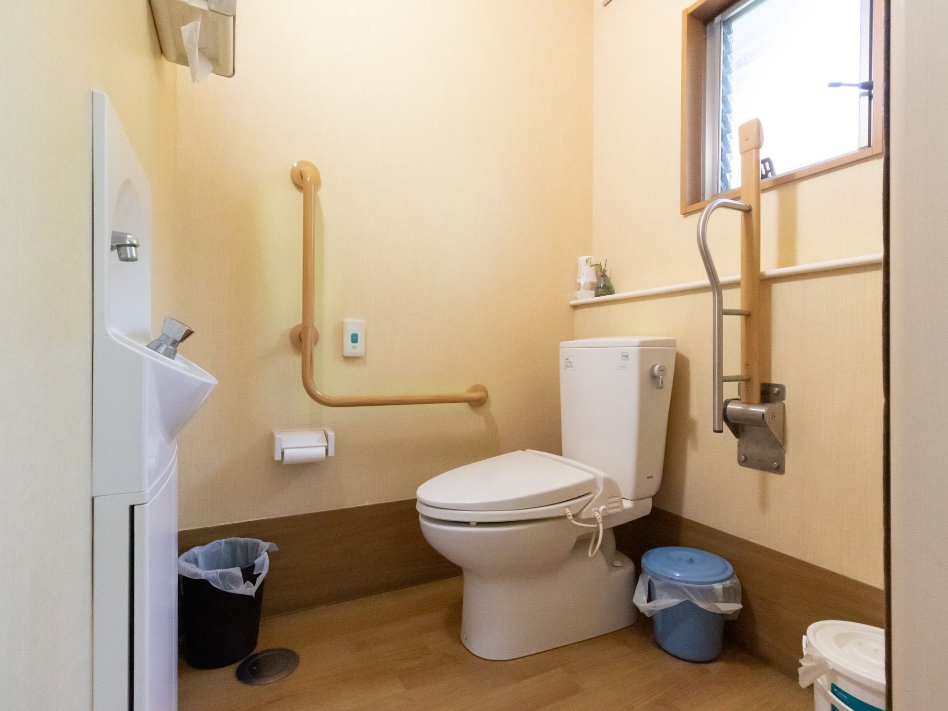 トイレの個室。壁には手すりと緊急用ブザー、手洗い場、ペーパータオルなどが取り付けられている。また床には複数のゴミ箱が置かれている。