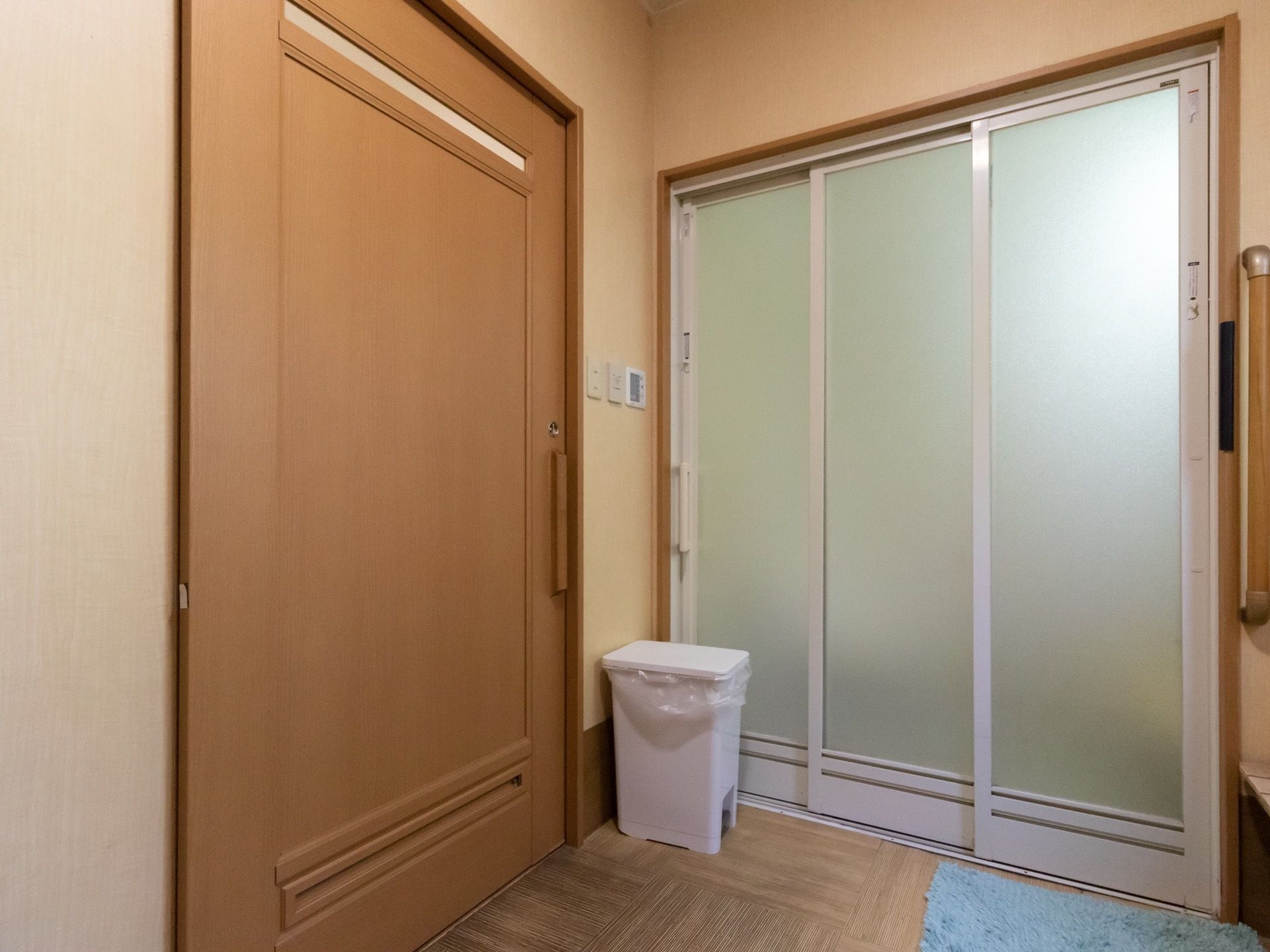 左に木目模様の引き戸の扉があり、右には浴室へと続く、スライド式の扉がある。床にはゴミ箱が置かれ、水色のバスマットも敷かれている。