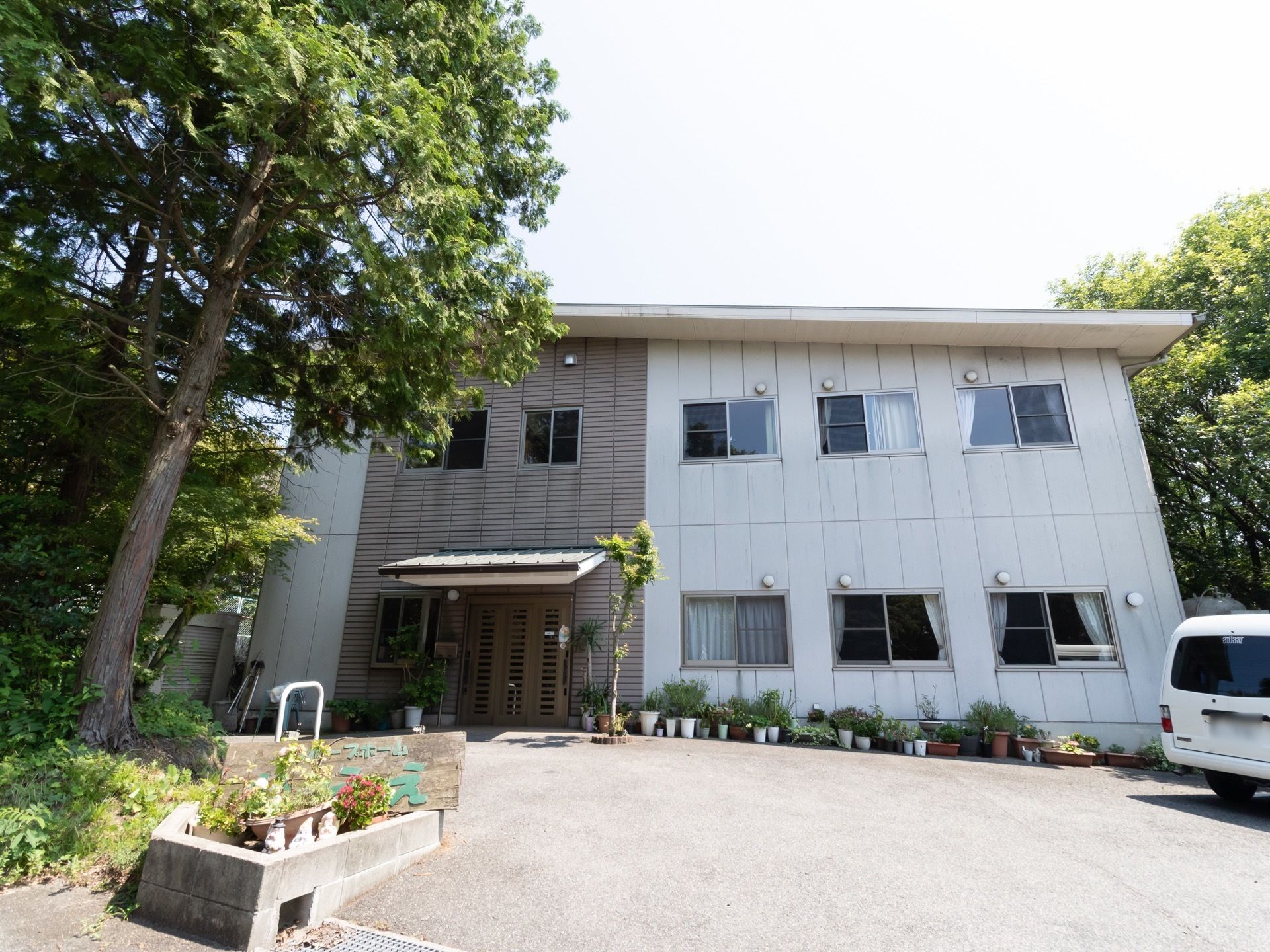 木々に囲まれた２階建ての建物があり、周囲には多数の鉢植えの植物が置かれている。前面には駐車場があり、車が停められている。
