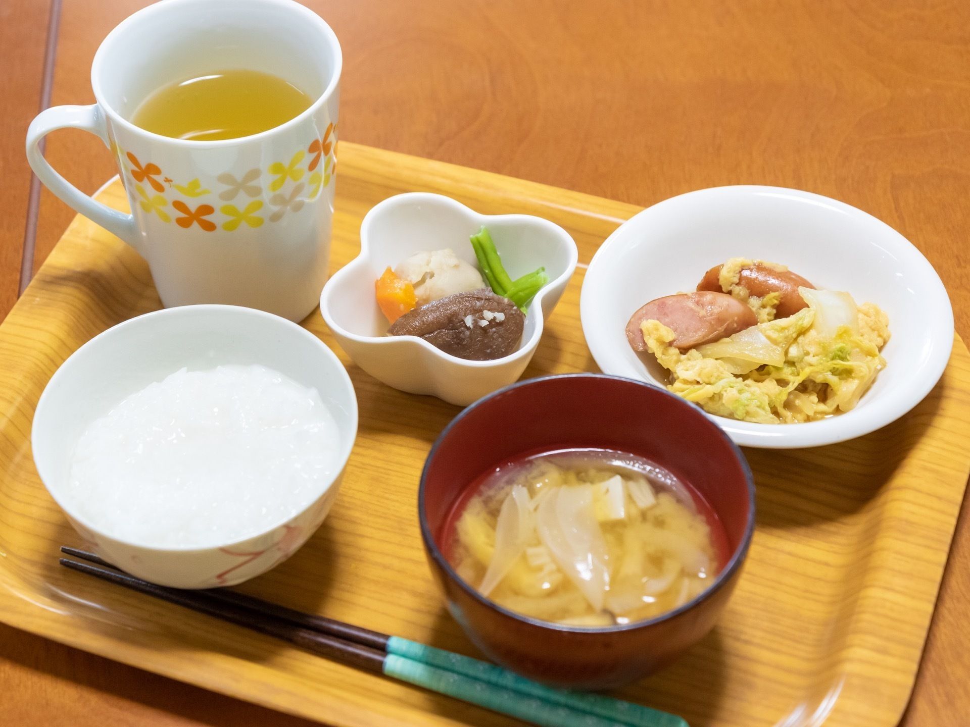 お茶とお粥、味噌汁、煮物、ソーセージと野菜などが入った皿がトレイの上に載せられ、箸が１膳添えられている。