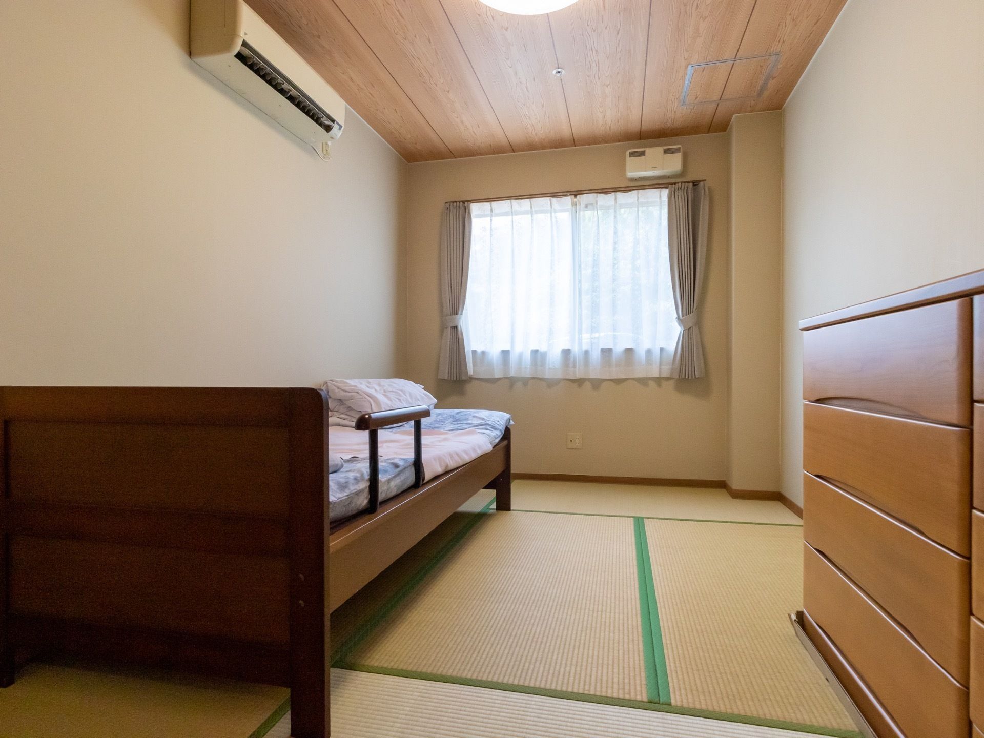 畳が敷かれた和室タイプの居室である。ベッドやエアコン、整理タンスが備わっている。窓にはカーテンがかけられている。