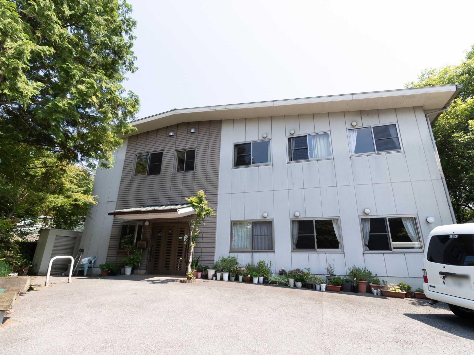 白を基調とした２階建ての施設で、敷地内に駐車場が完備されている。外壁沿いには、鉢植えが複数置かれている。