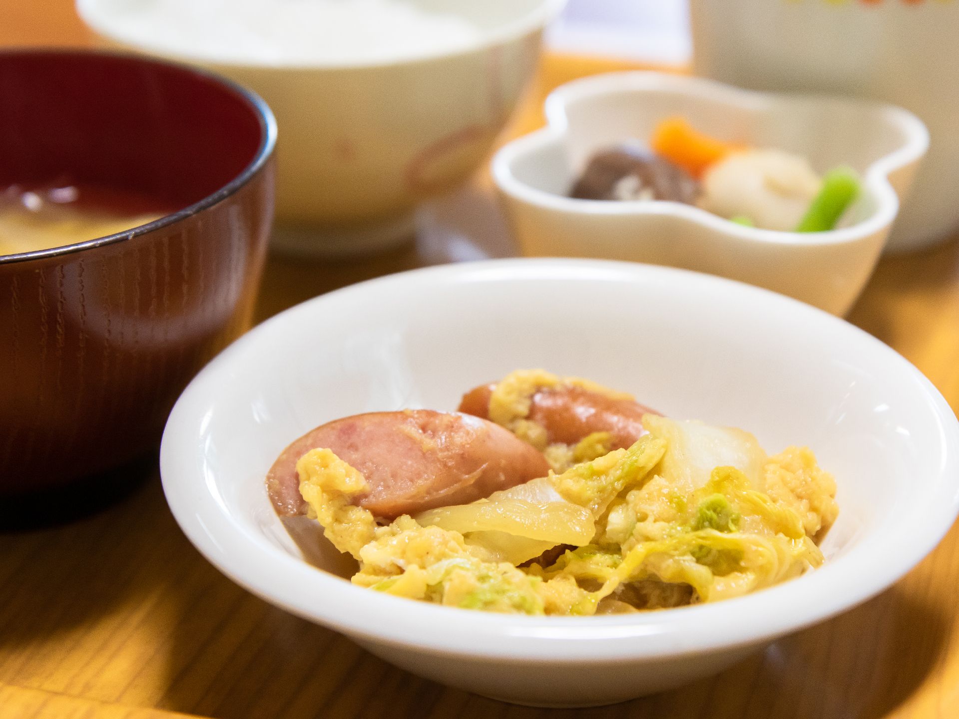 ウィンナーとスクランブルエッグの盛られた白い皿、味噌汁や小鉢が添えられた食事例