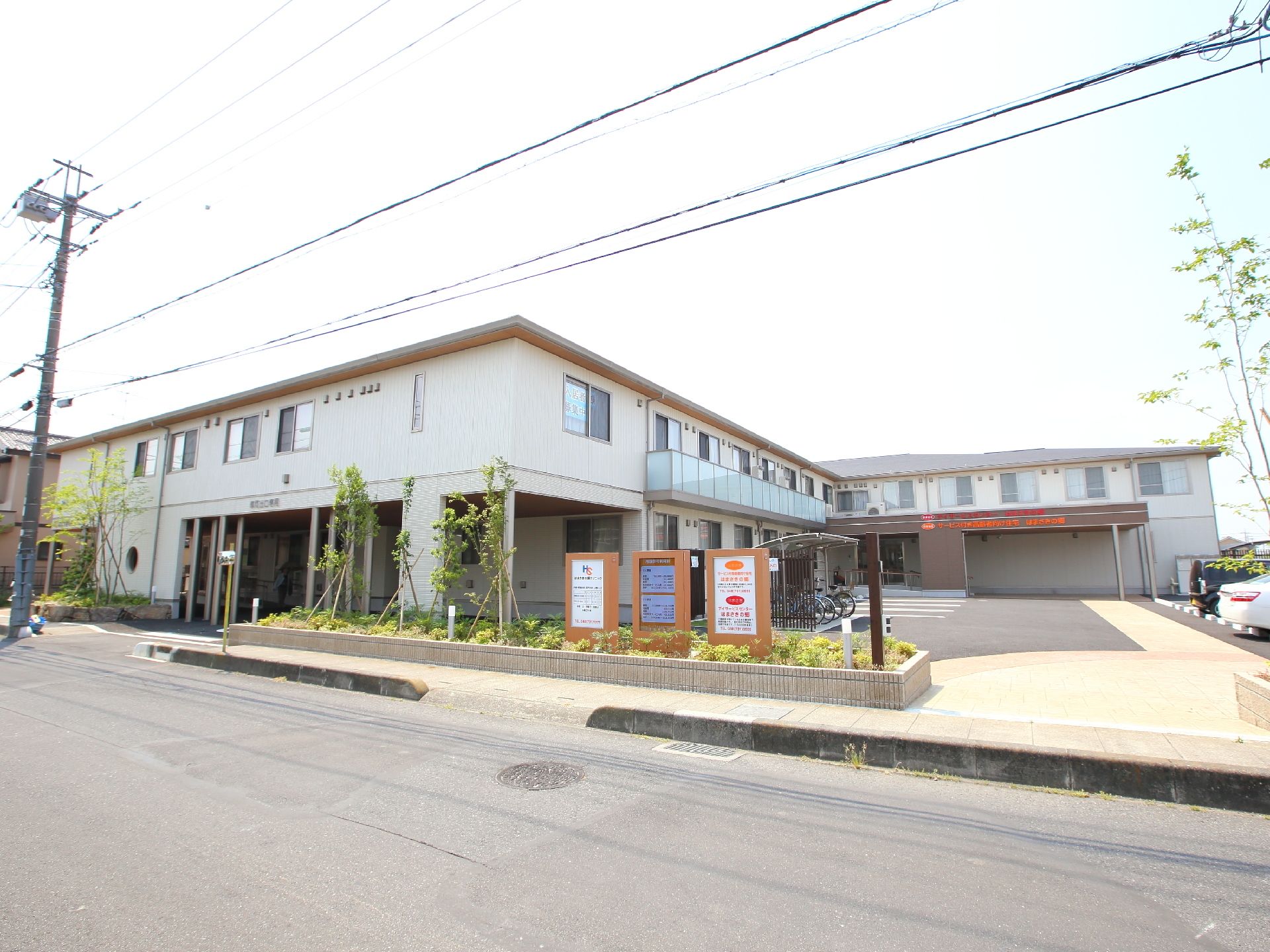 手前の道路からの入口に看板があり、その向こうにある白い外壁でL字型の2階建ての建物