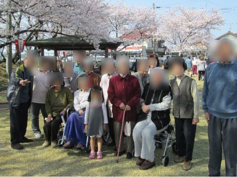 満開の桜が後ろに並び、東屋のあるスペースで車椅子に座ったりその後ろに立ったりして集合写真におさまる高齢者