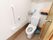 蓋の付いていない洋式水洗トイレが設置してある。右側の壁に手すりがあり、操作ボタンやトイレットペーパーが2つある。呼び出しボタンもある。