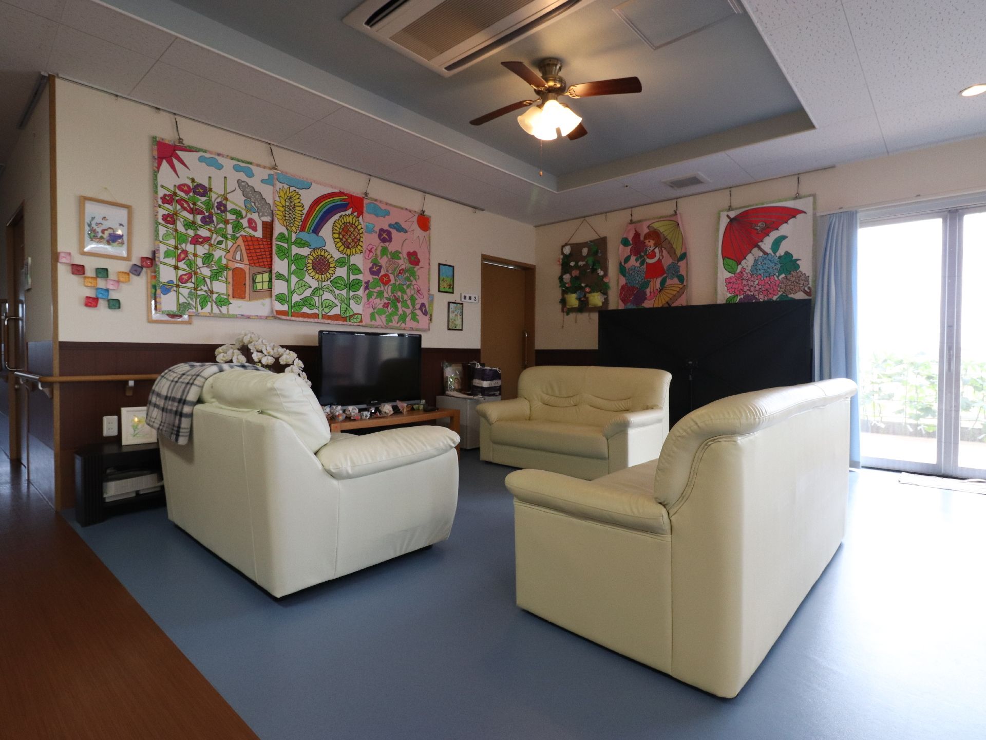 白いソファーがテレビの前にある。壁にはひまわりやアジサイなどの絵があり季節感を感じる。天井にはシーリングファンが付いている。
