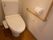 白地の壁とフローリングの床面が組み合わさった落ち着いた雰囲気のトイレである。洗浄等の操作ができるボタンは便座の横に付属している。