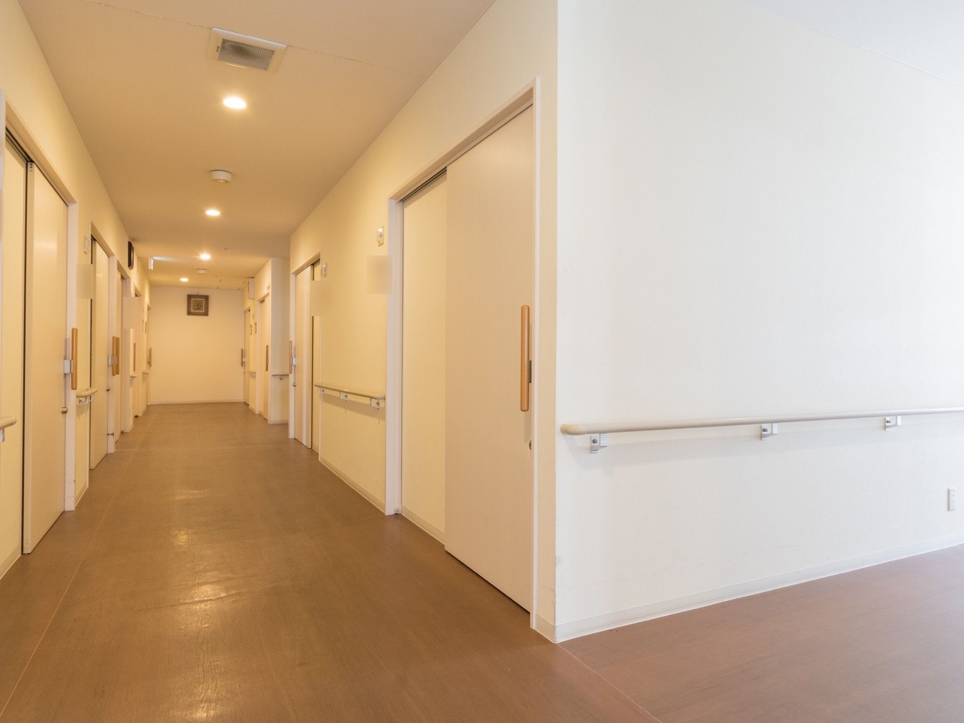 通路の床は茶色く、壁は白色である。そこに白い部屋の扉が複数並んでいる。壁には手すりが取付けられている。