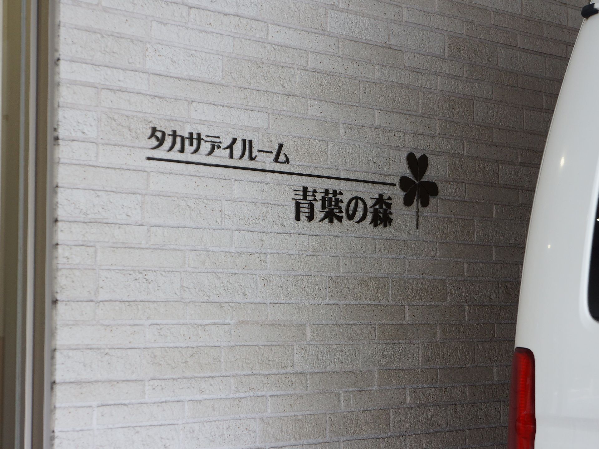 白い外壁に施設名が直接書かれていてクローバーのデザインもある。その場は駐車場になっているのか白い車の後ろ部分が映っている。
