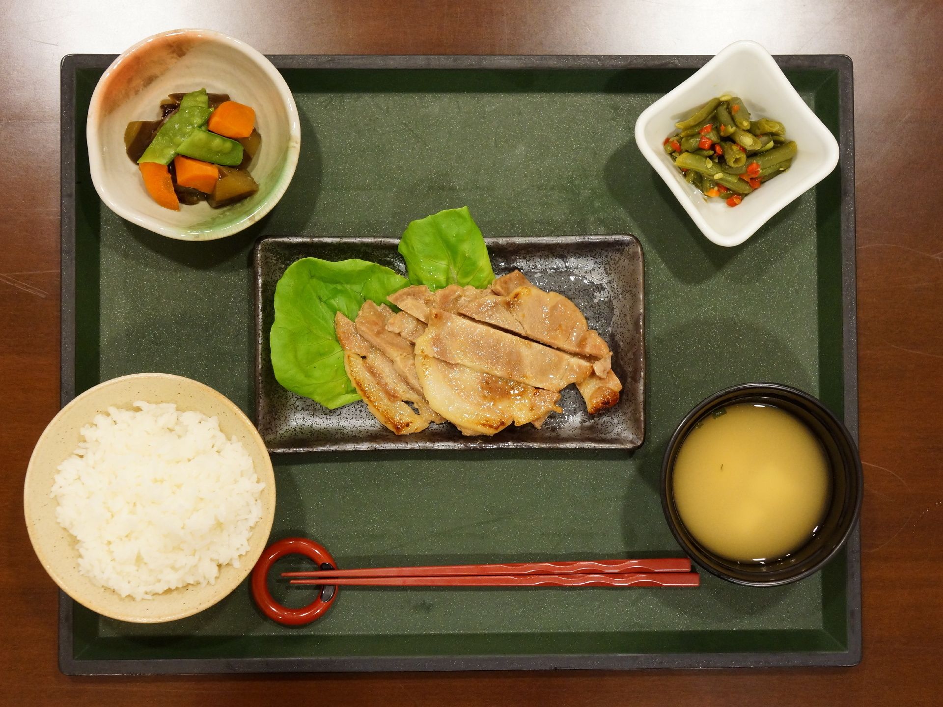 グリーンの四角いお盆の上に食事が乗ってる。メインは長方形のお皿に盛りつけられた豚肉料理になっている。他に野菜料理の入った小鉢がある。