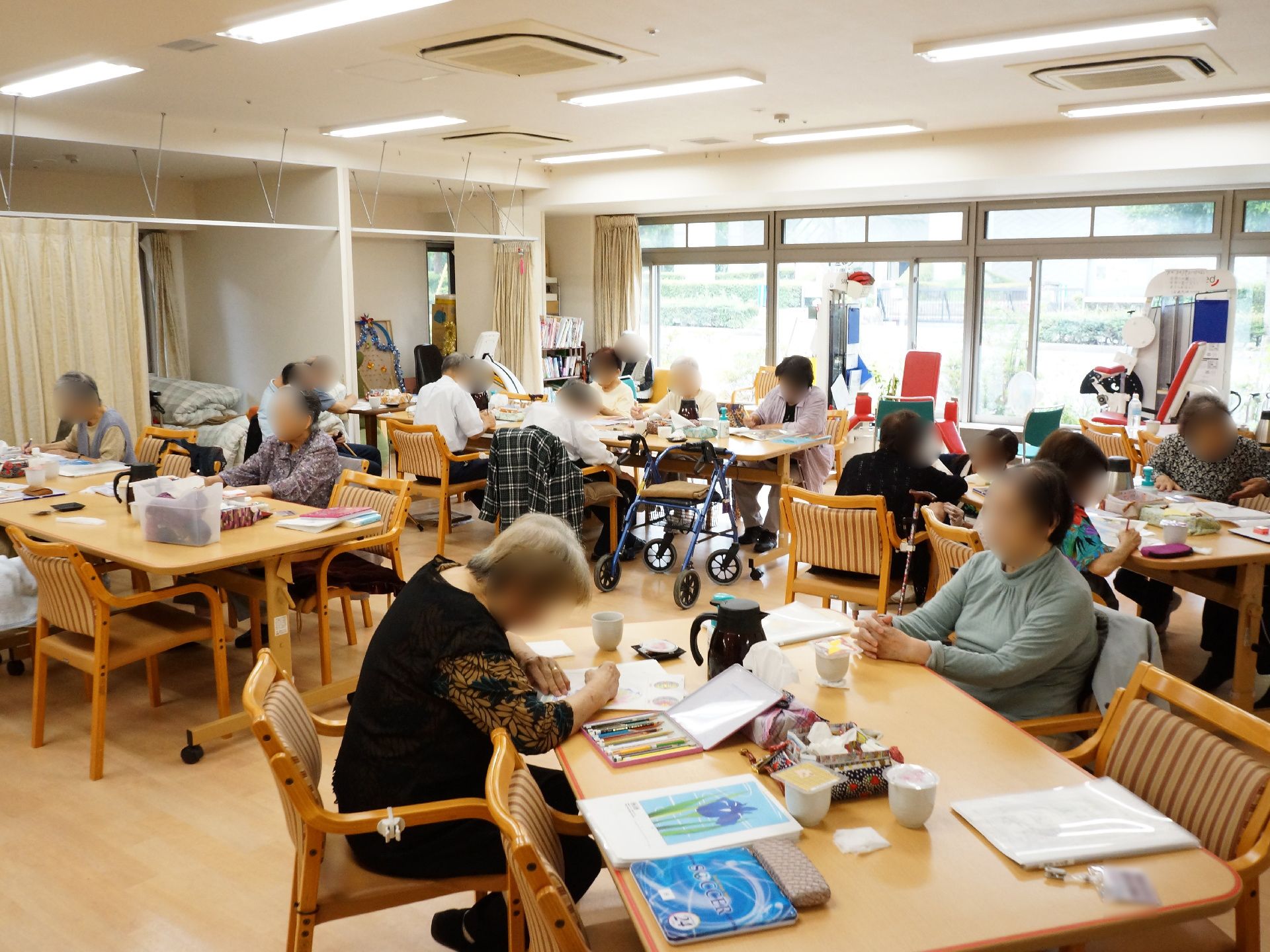 ガラスのドアが壁一面にあり陽が明るく差し込んでいる。多くの入居者様がそれぞれに好きなことをしている。窓際にはトレーニングマシンがある。