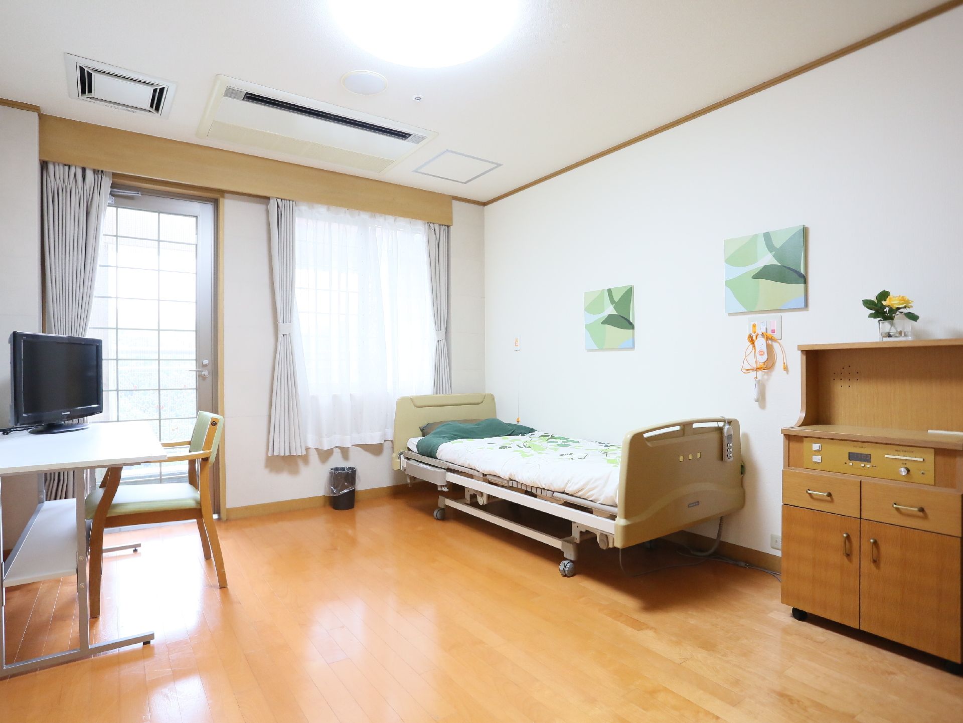 正面に掃き出し窓と腰高窓がある。向かって右側には低床型のベッドがある。間を空けて反対側の壁にはテレビが並んでいる。