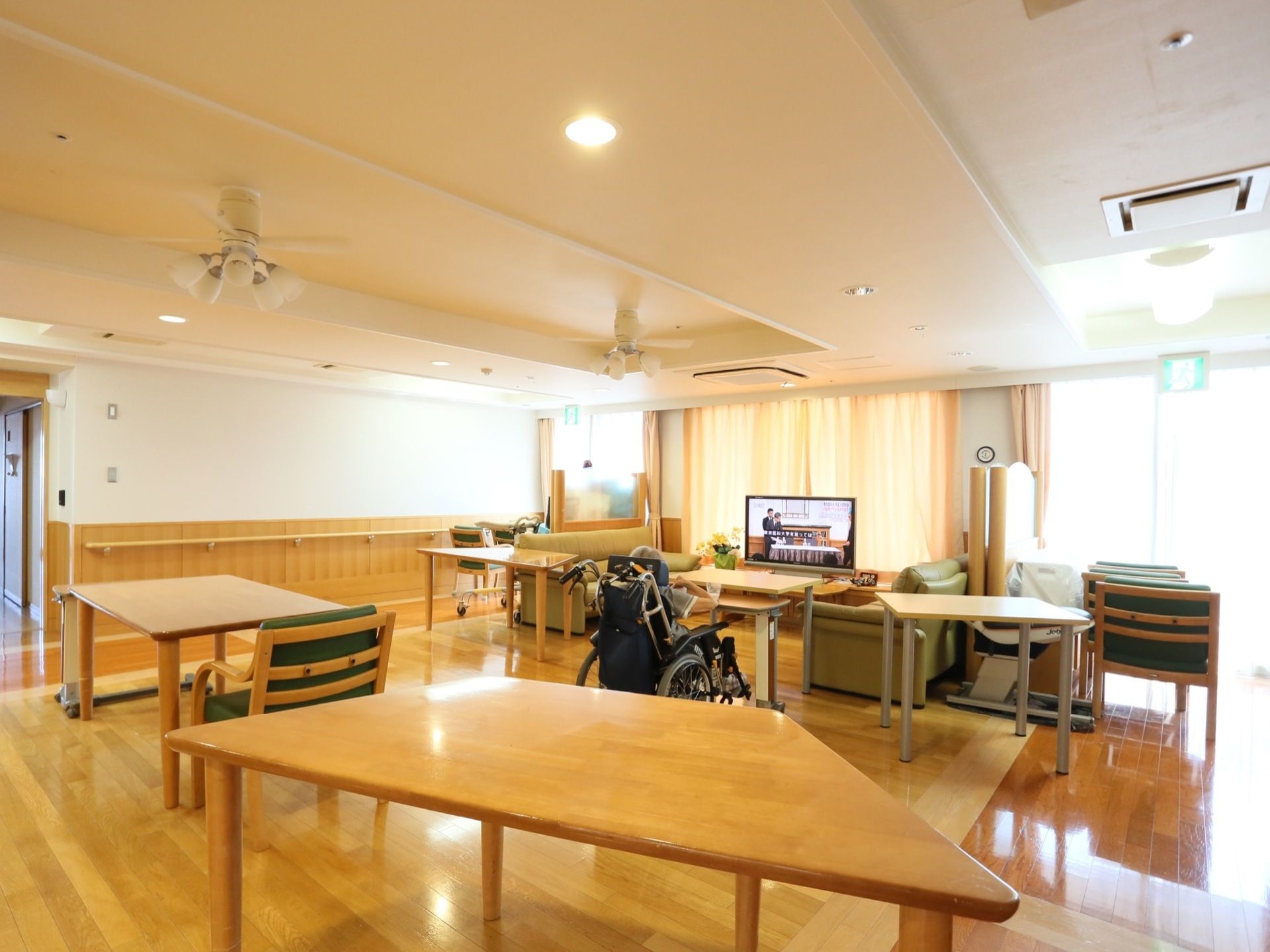 広々とした共有スペースで入居者様がテレビを鑑賞しながらくつろいでいる。壁側には手すりも備わっている。
