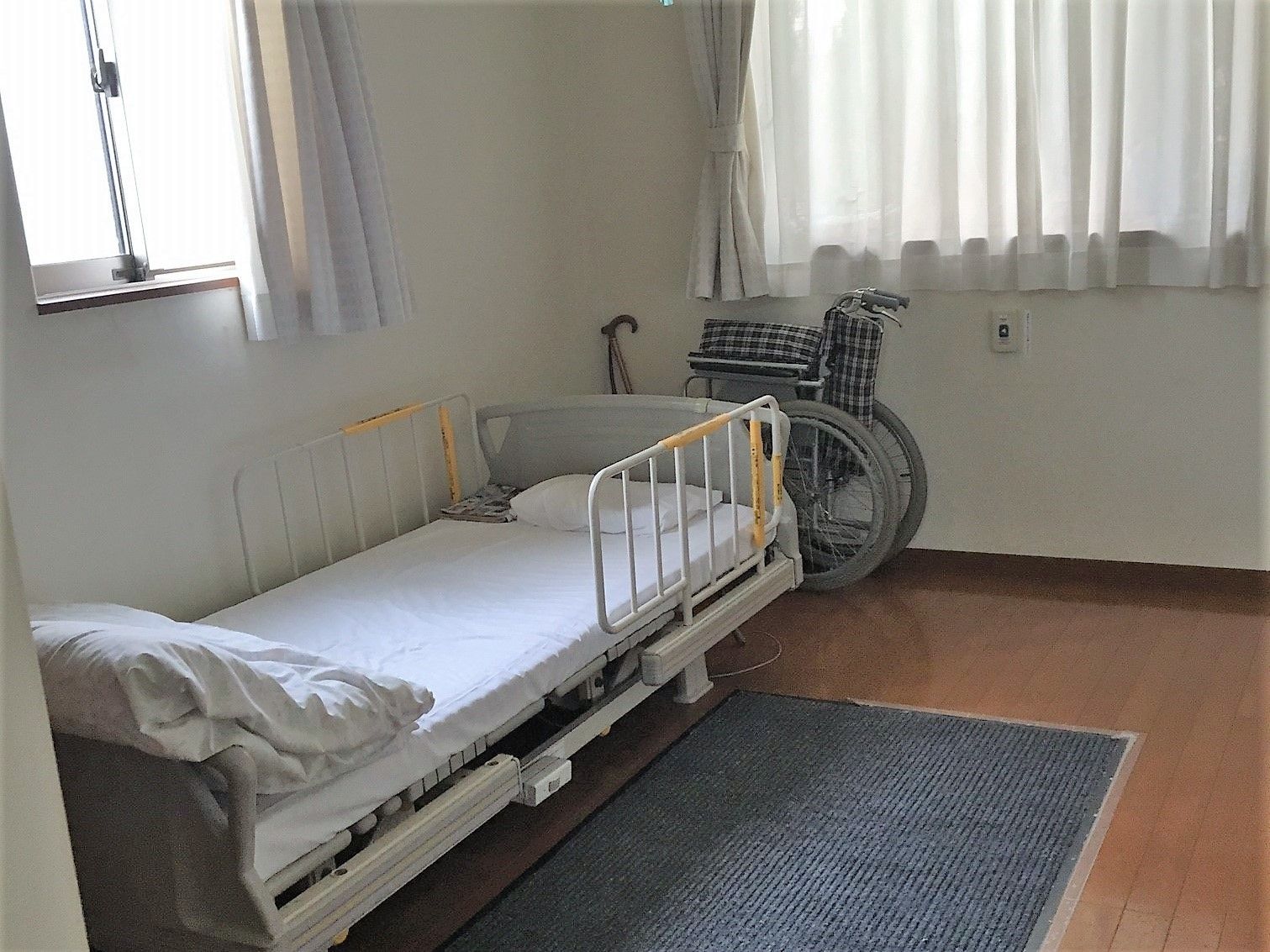 正面と横側に窓があり、自然光が降り注ぐ明るい空間の居室である。壁際に介護用ベッドを備え、横に車椅子を置いている。