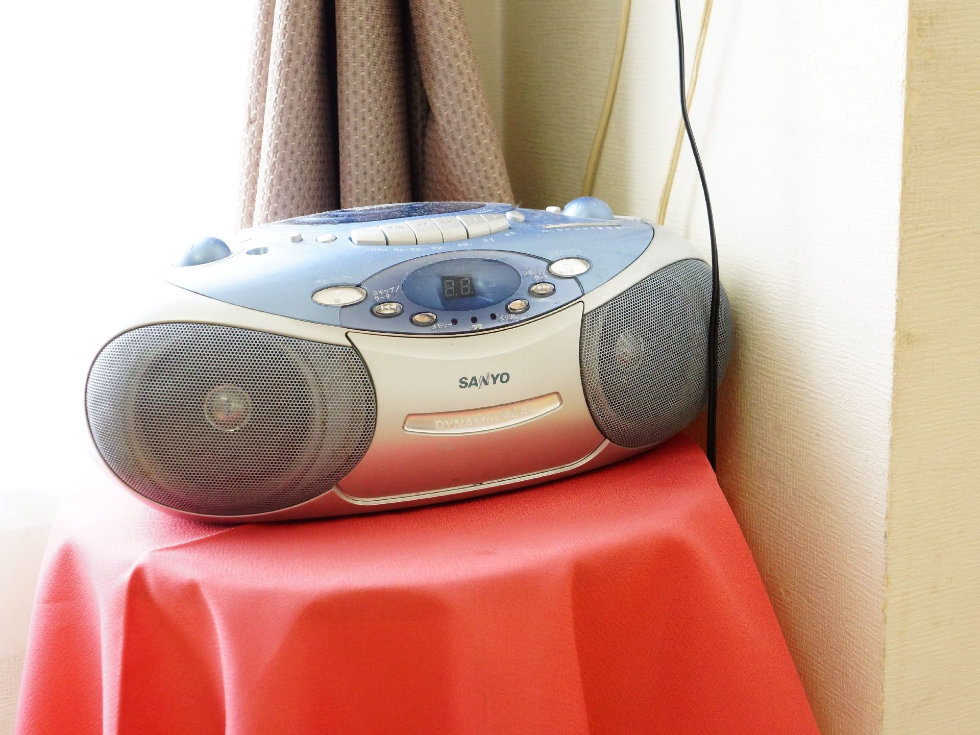 光の差し込む窓にカーテンがかかっており、手前の台に赤いクロスがかけられ上にはラジカセが置かれている。