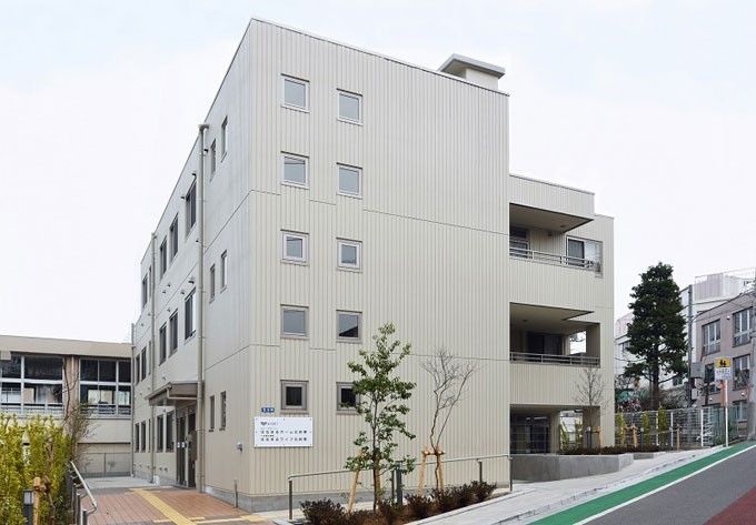 落ち着いたホワイト系の３階建て施設である。広いスロープが設置されており、中央に黄色い点字ブロックが配置されている。