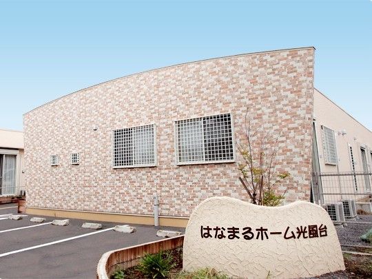 建物の外壁はベージュ系のレンガ調で、上部が緩やかな曲線を描いており、美術館のような外観である。建物前には駐車場や花壇、施設名の看板がある。