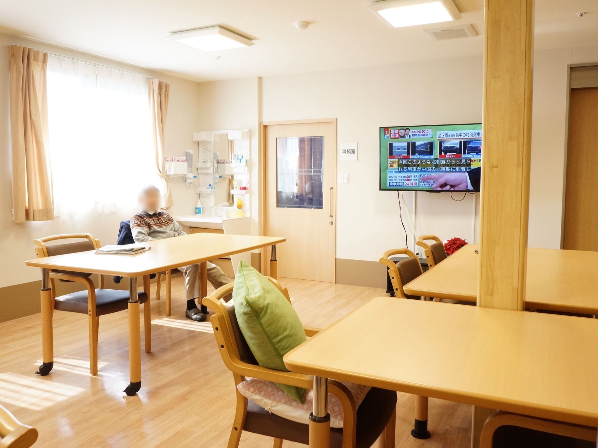 大きなテレビの設置された食堂。角には洗面台があり、隣に入居者様が座っている。４人がけのテーブルが数個並べられている。