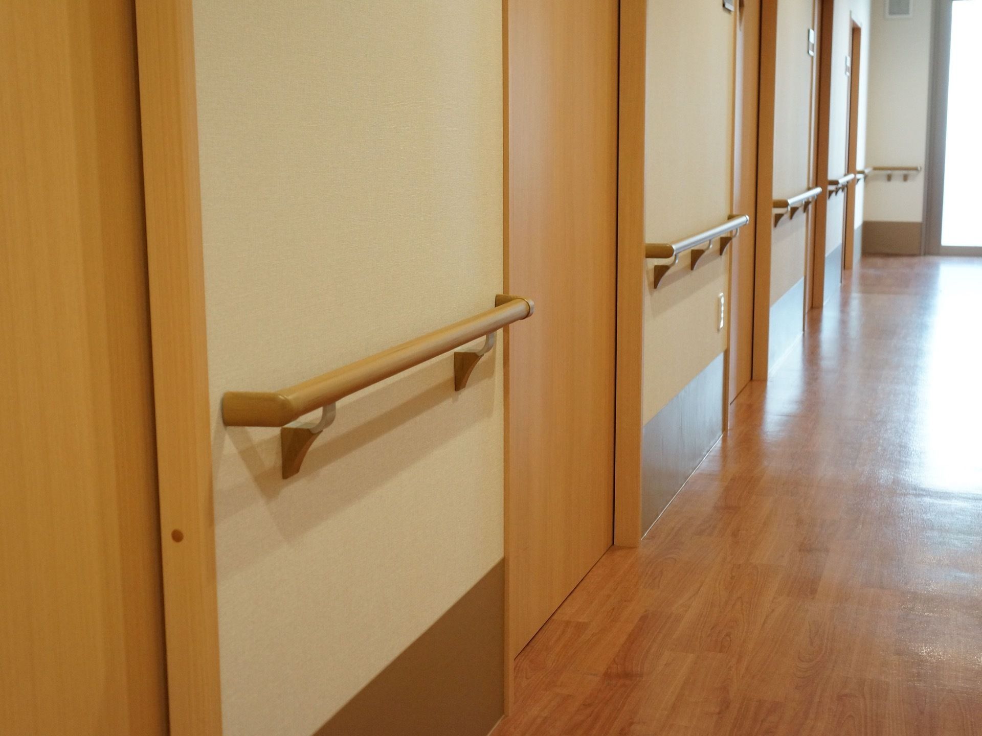 直線に伸びた廊下には、扉以外の壁全てに手すりが設置されている。床と扉はナチュラルな木目柄。廊下の突き当たりには窓がある。