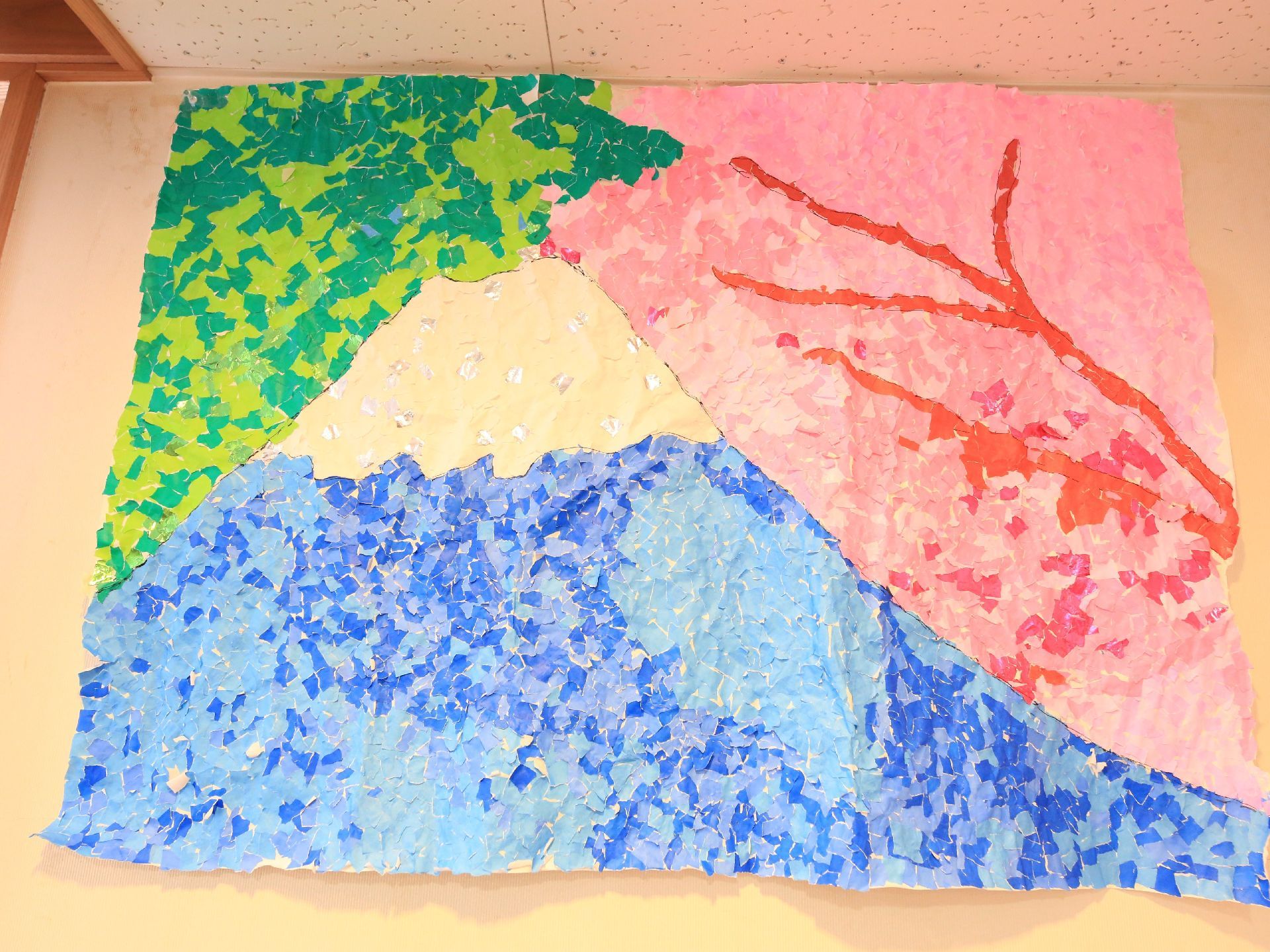 ちぎり絵の作品が壁に飾られている。色とりどりの細かな紙片を組み合わせて、雪をかぶった山やピンク色の花、緑が表現されている。