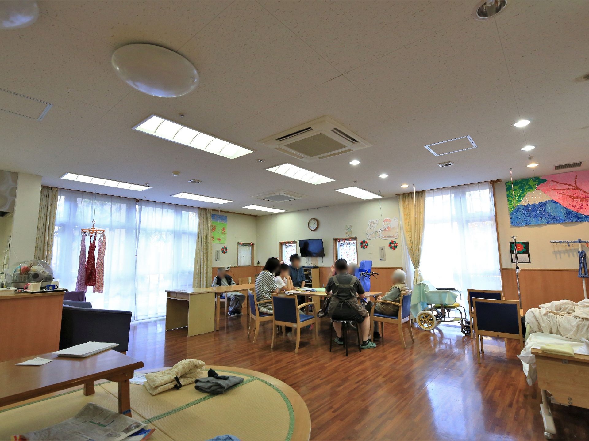 共有スペースのテーブルに、入居者様とスタッフ数人が集まっている。壁の天井近くには、大きなちぎり絵の作品が飾られている。