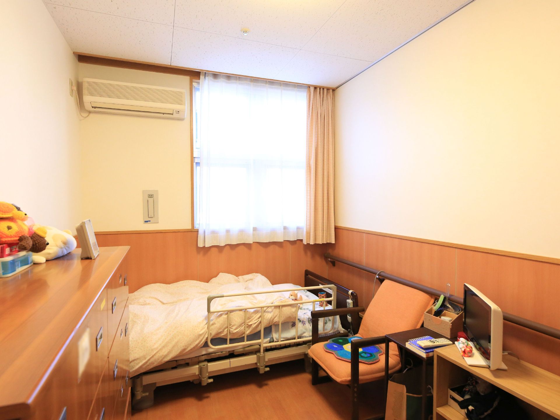 居室には転落防止柵つきのベッドが設置されている。窓の横にはエアコンがあり、壁際には収納タンスが置かれている。