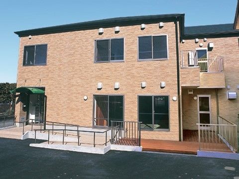 ２階建てのブラウンを基調とした建物。緑色の屋根が付いた玄関の前には、手すりが付いた階段やスロープが設置されている。