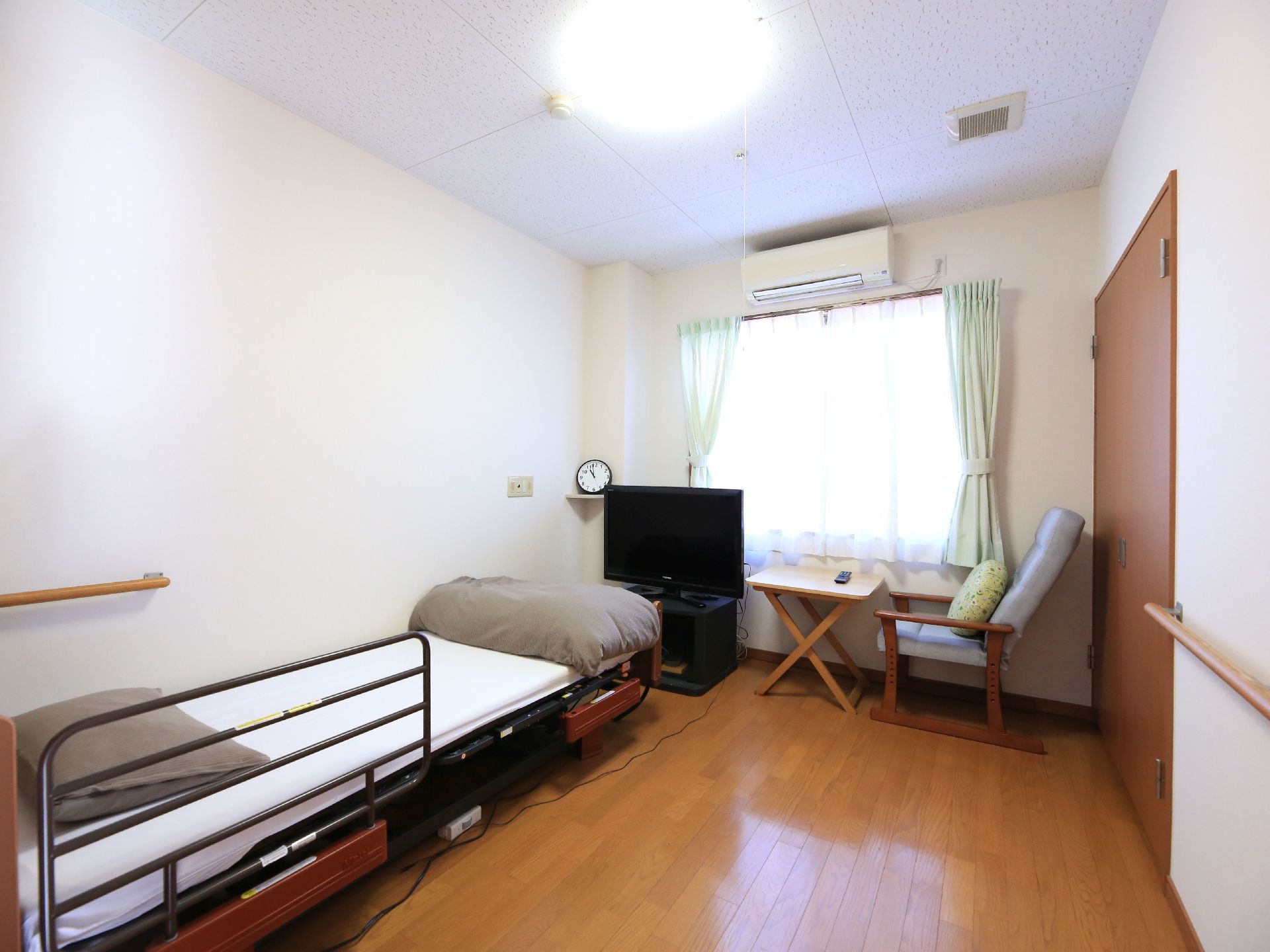 居室には、転落防止柵つきの介護ベッドが設置されている。窓の上部にはエアコンがあり、窓際にはテレビが置かれている。