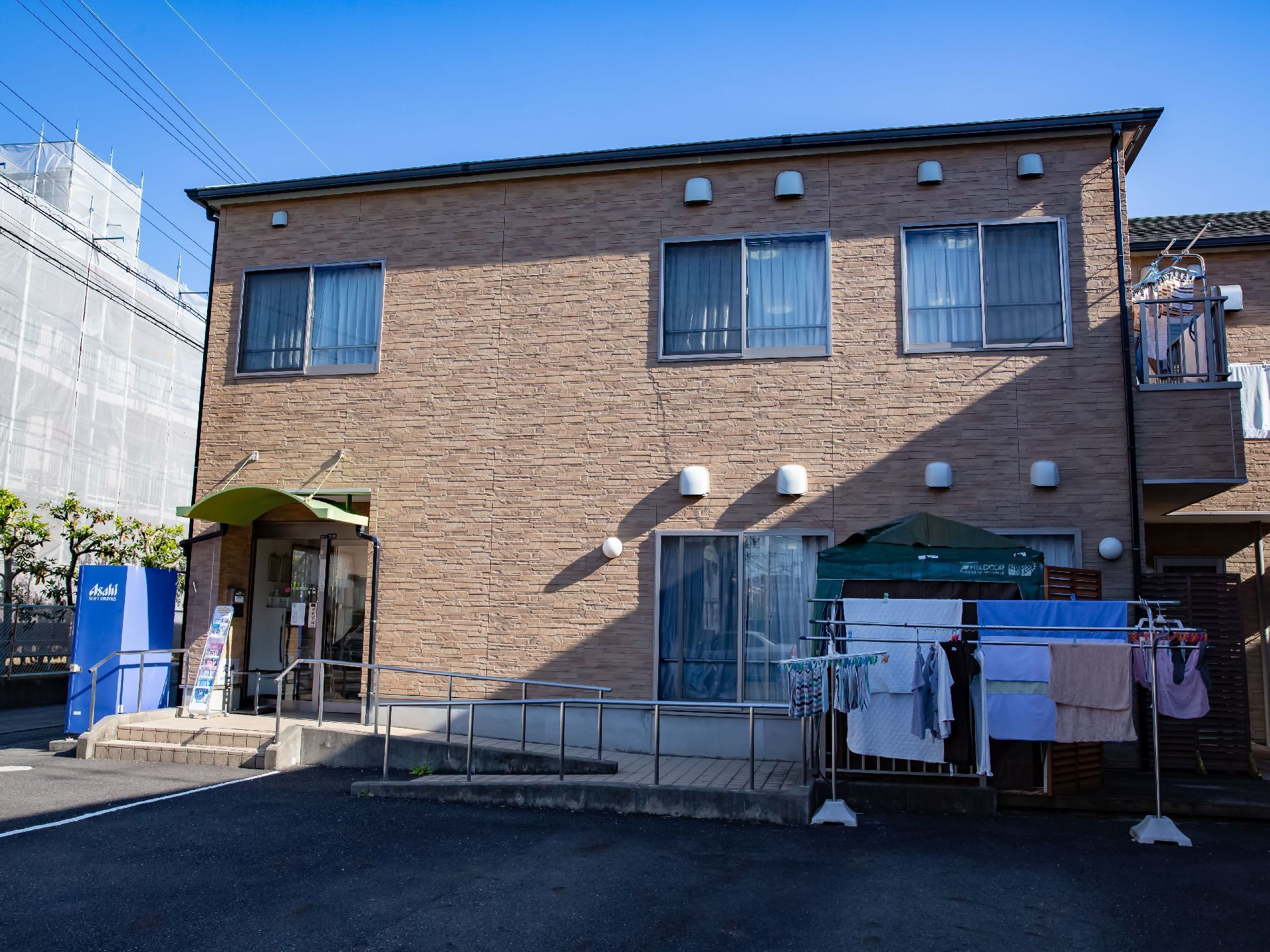 石積み状の模様の外壁の２階建ての施設である。２階のベランダと敷地内に洗濯物が干されている。玄関脇に飲料の自動販売機がある。
